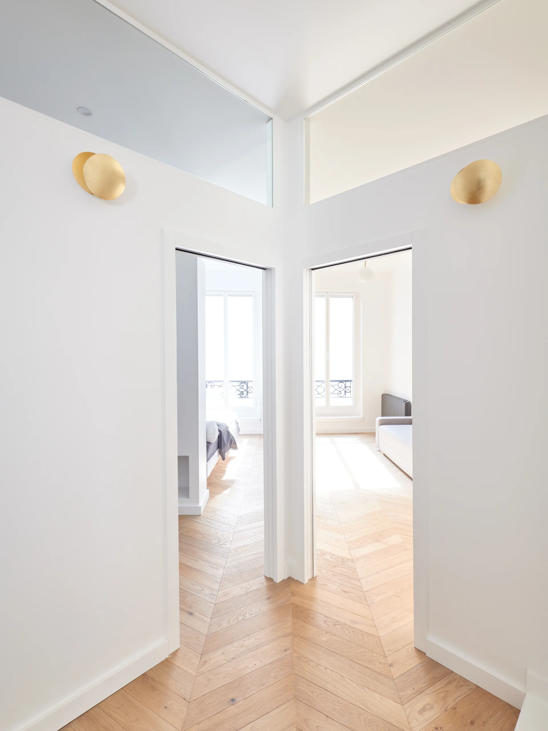 A bright white hallway features light wood herringbone flooring, two open doorways with glass transom windows, and circular gold wall sconces.