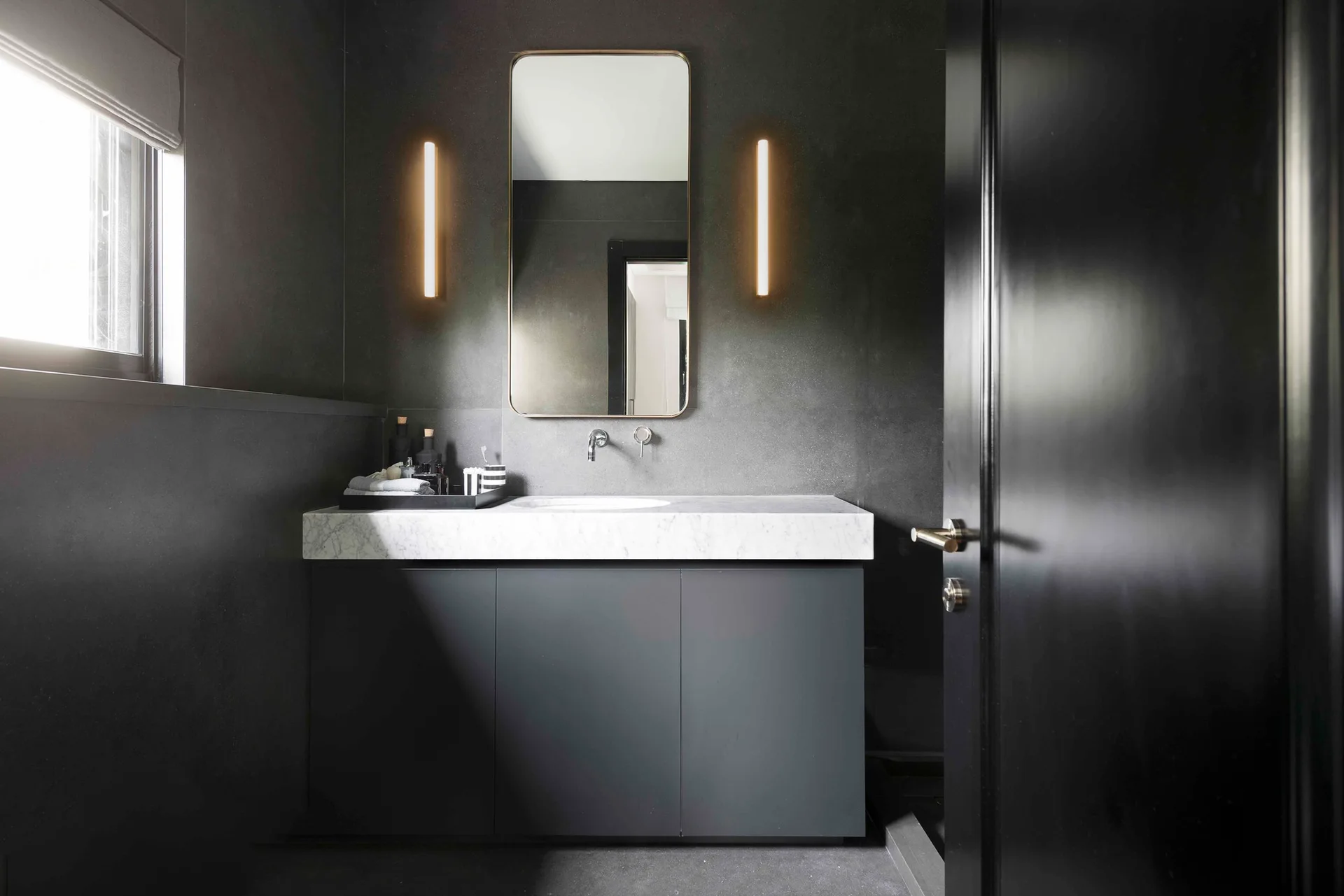 A dark gray bathroom featuring a black cabinet vanity with a white marble countertop, a rectangular mirror, and two glowing vertical wall sconces.