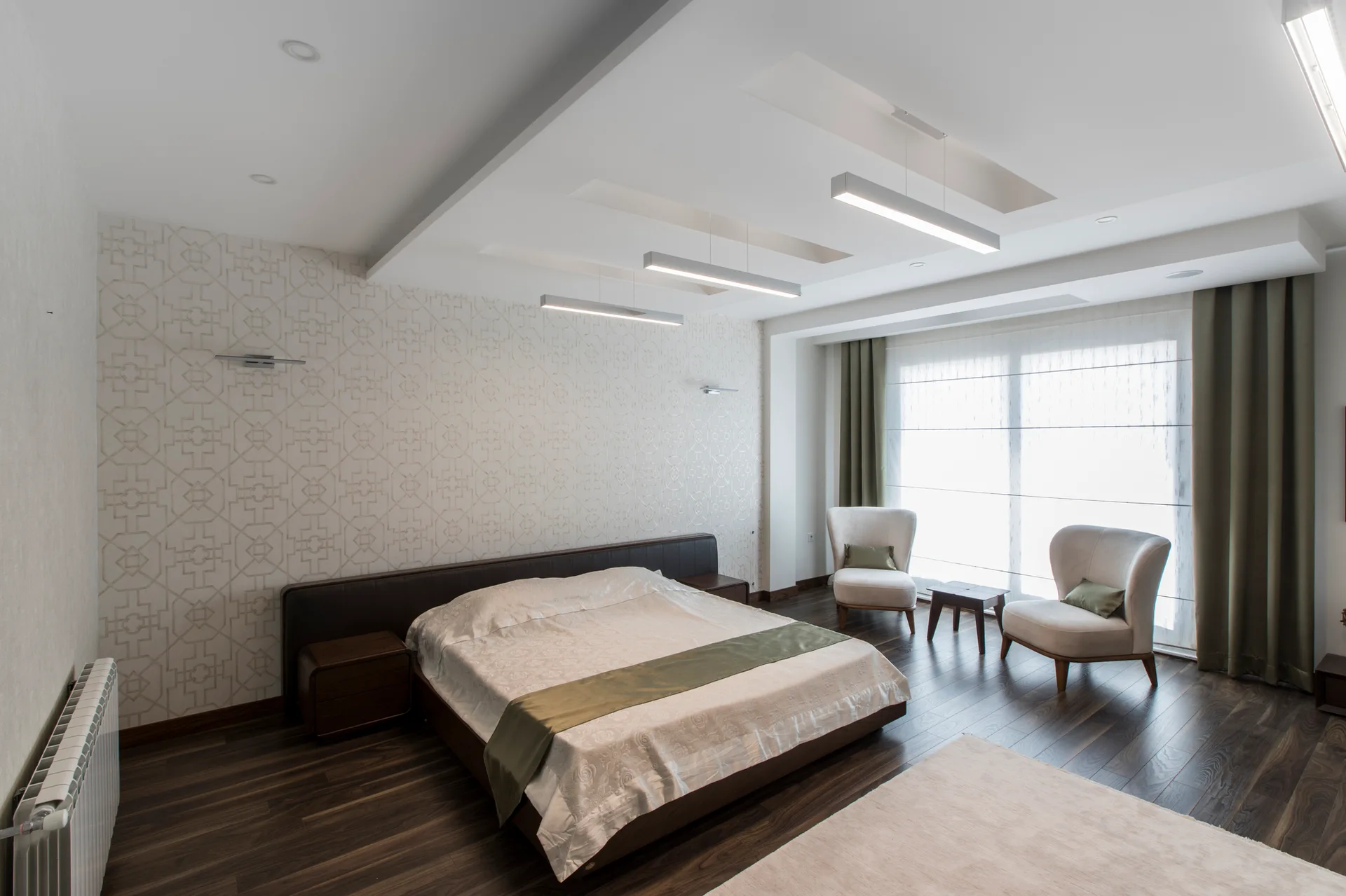 A contemporary bedroom features a low bed against geometric wallpaper, dark wood floors, and two light armchairs by a large window.