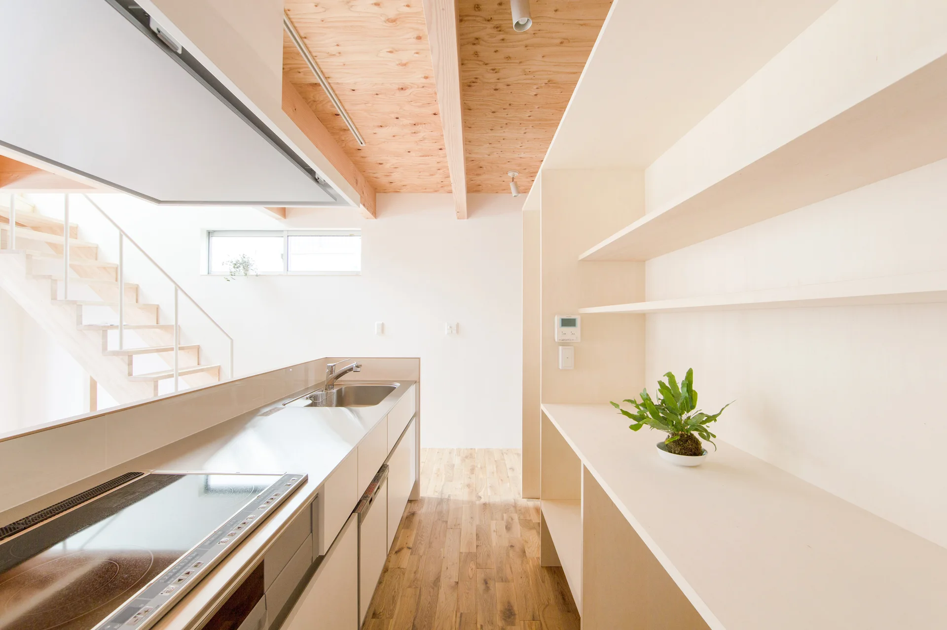 A minimalist kitchen features a light wood floor, white cabinets, open plywood shelving, and an exposed wood beam ceiling with a staircase in the background.