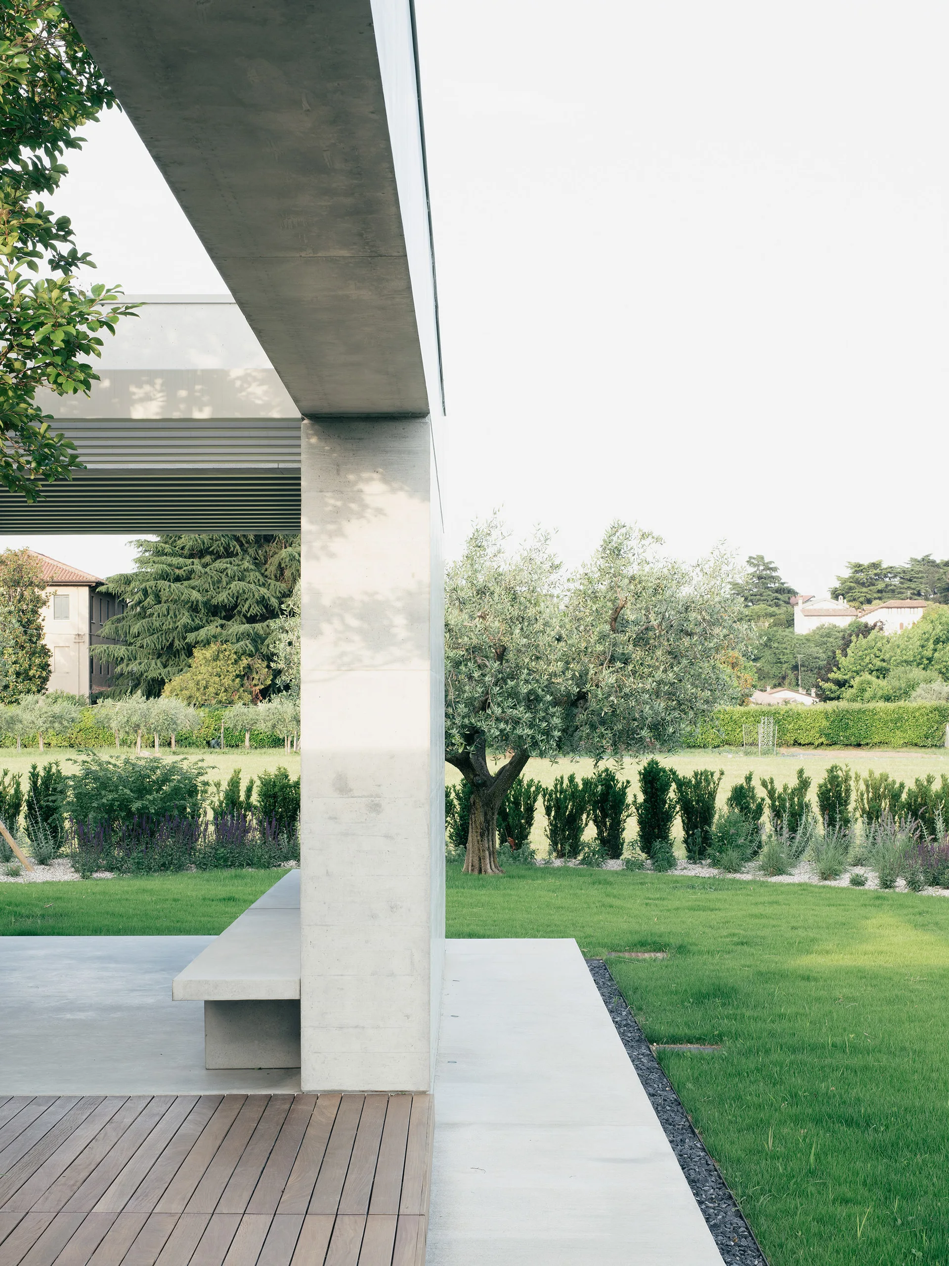 A minimalist concrete pavilion features a built-in bench, wooden plank decking, and an open view of a landscaped green lawn.