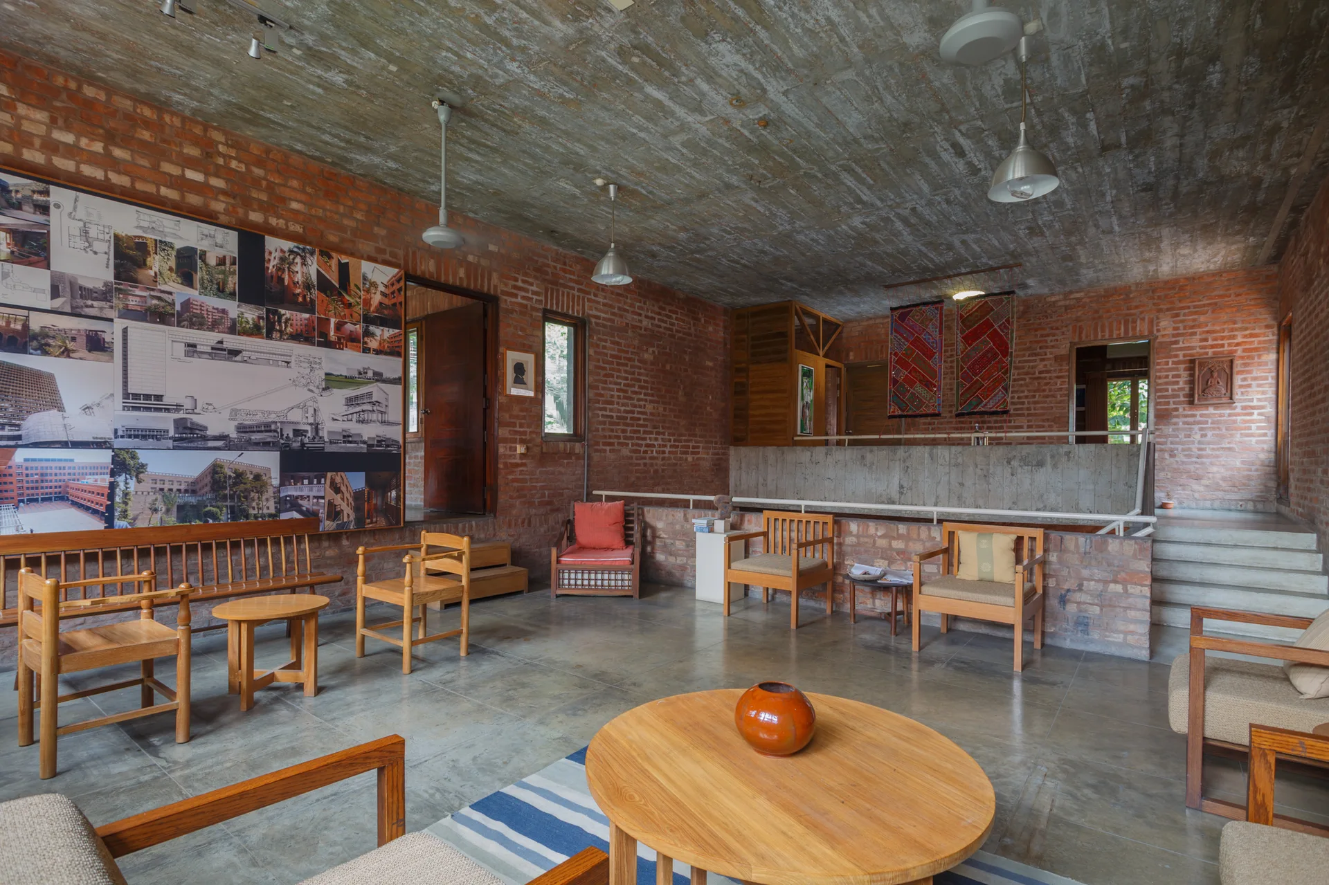 A spacious lounge featuring exposed red brick walls, a textured concrete ceiling, wooden armchairs, and a round coffee table.