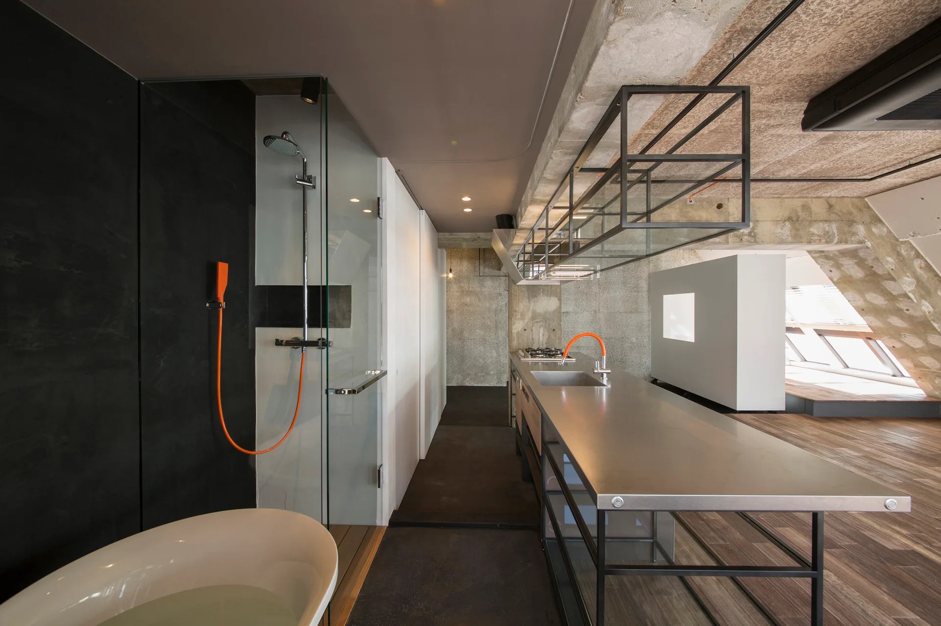 An open industrial loft features a stainless steel kitchen island and a glass-enclosed shower next to a freestanding bathtub under a concrete ceiling.