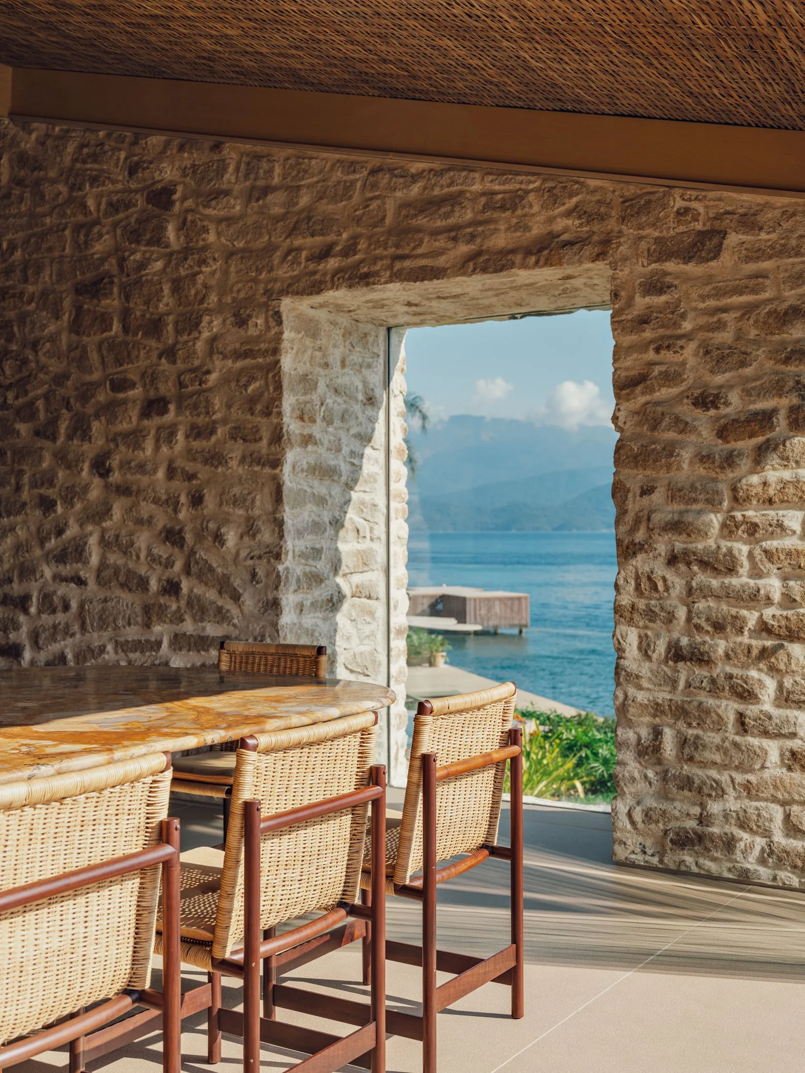 A dining area features a stone-topped table and woven wicker chairs next to a thick stone wall with a deep window opening to the sea.