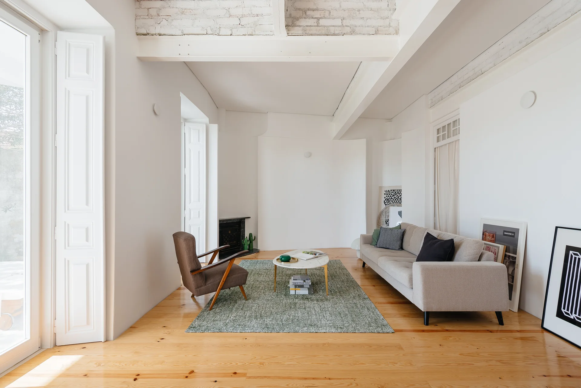 A bright living room features a grey sofa, a mid-century armchair, and a marble coffee table on a green textured rug under white exposed ceiling beams.
