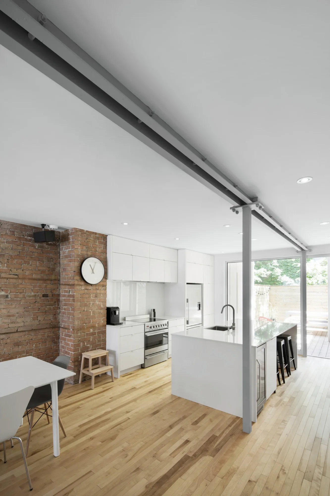 A modern kitchen featuring white minimalist cabinetry, a large white island with bar stools, a red brick pillar, and a grey structural steel beam.