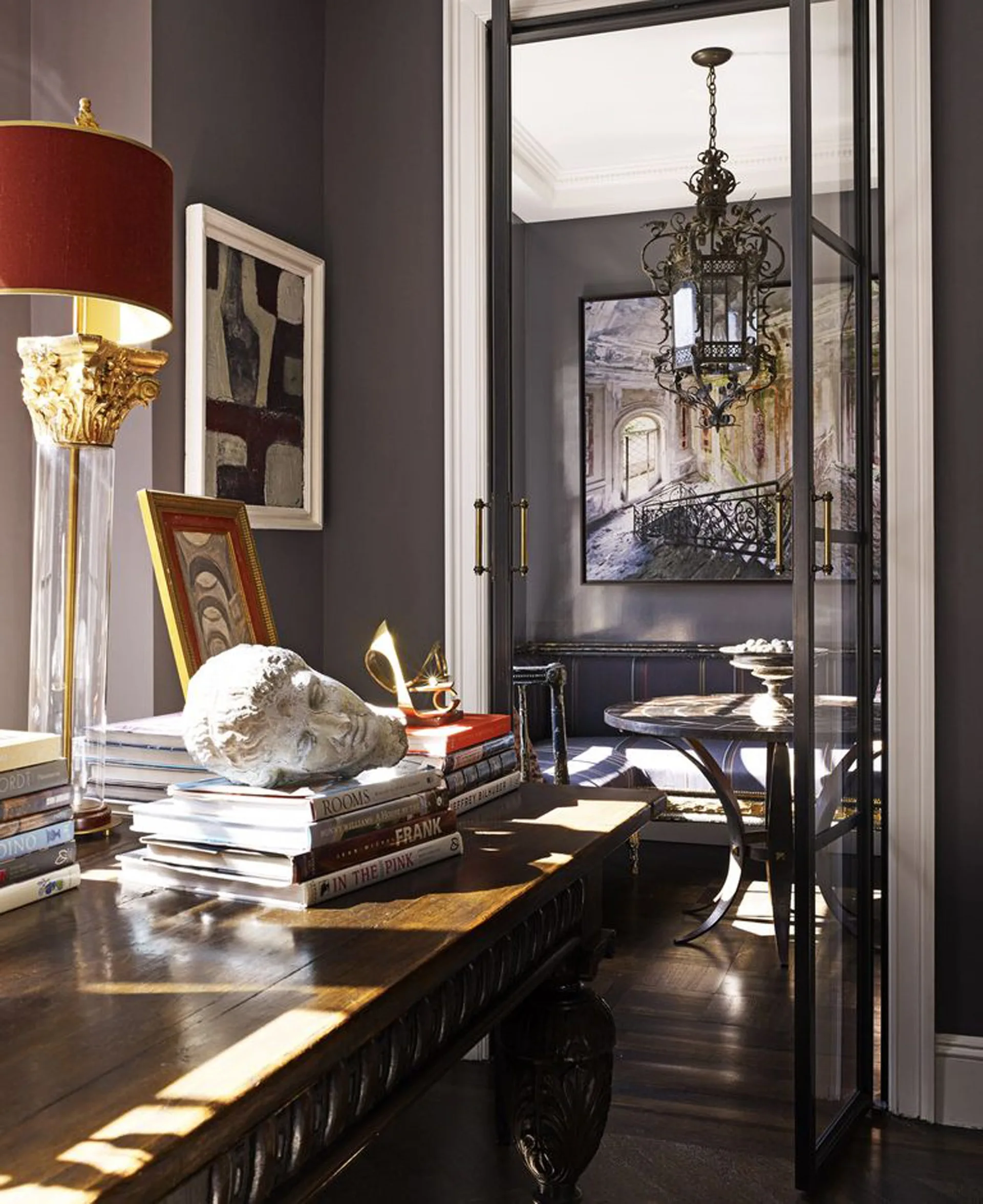 A dark gray room features a wooden console table with stacked books and a sculpture, looking through black-framed glass french doors to a seating area.