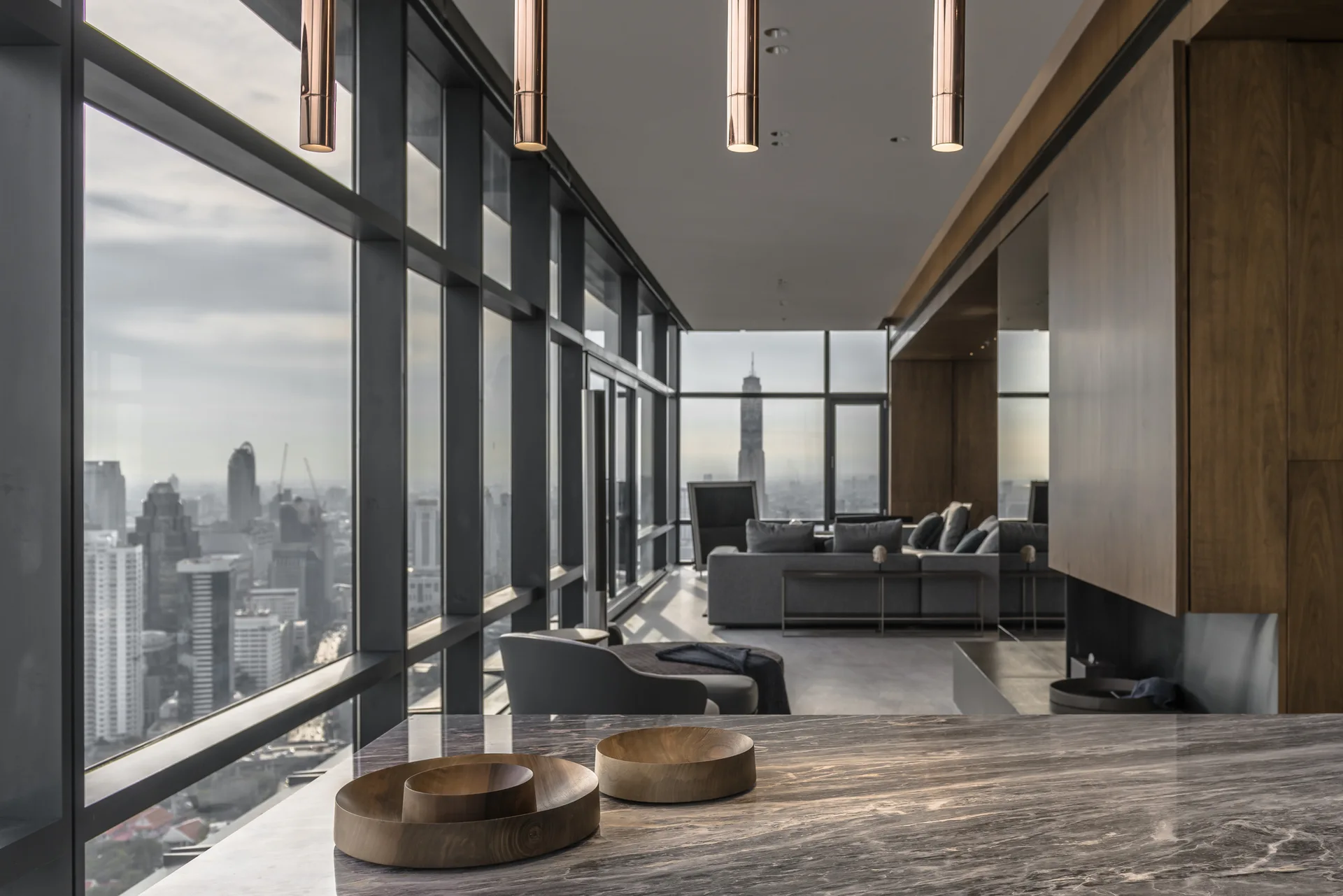 A veined stone countertop with wooden bowls sits in the foreground of a modern high-rise living area with floor-to-ceiling windows and a gray sofa.