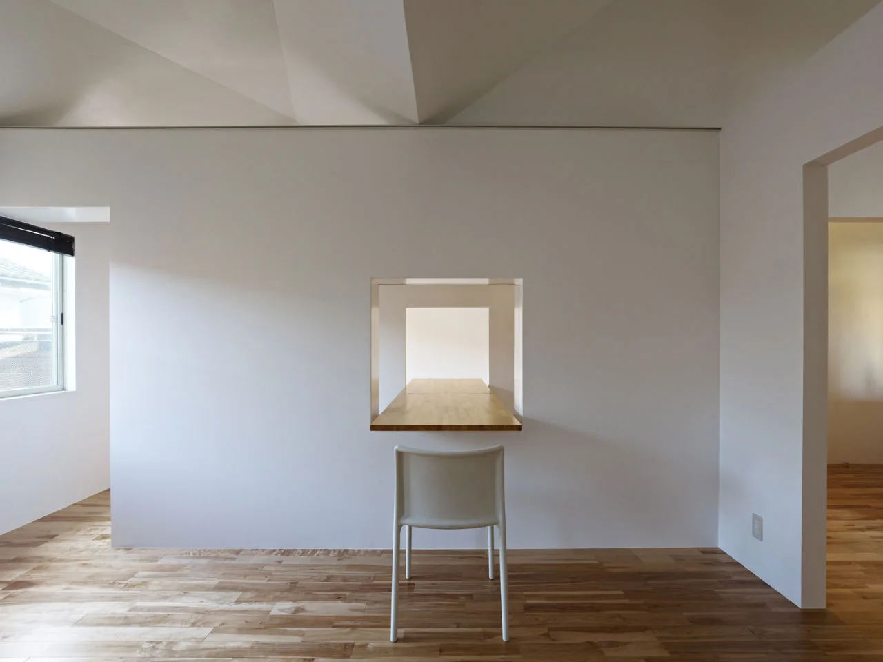 A minimalist white room with a wooden desk built into a square wall opening and a single white chair on a natural wood floor.