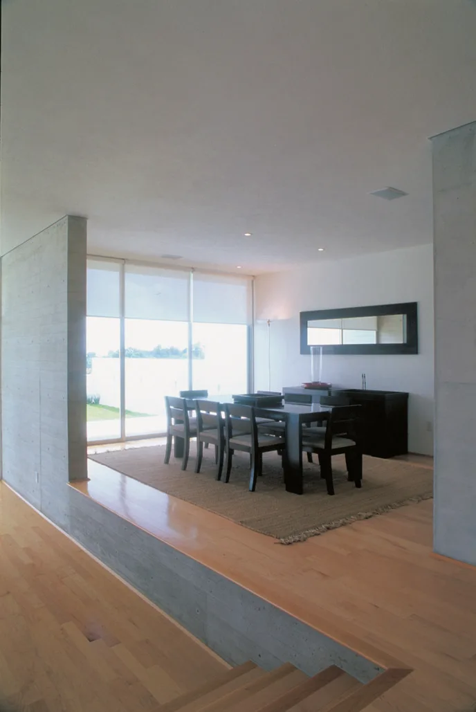 A sunken dining room featuring a dark wood table and chairs on a woven rug, flanked by exposed concrete walls and light wood flooring.