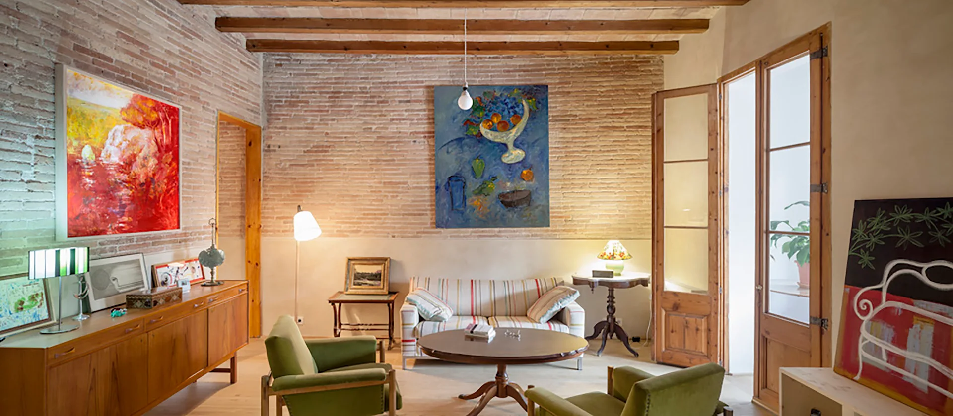 An eclectic living room with a brick wall, wood ceiling beams, a striped sofa, green armchairs, and large colorful paintings.