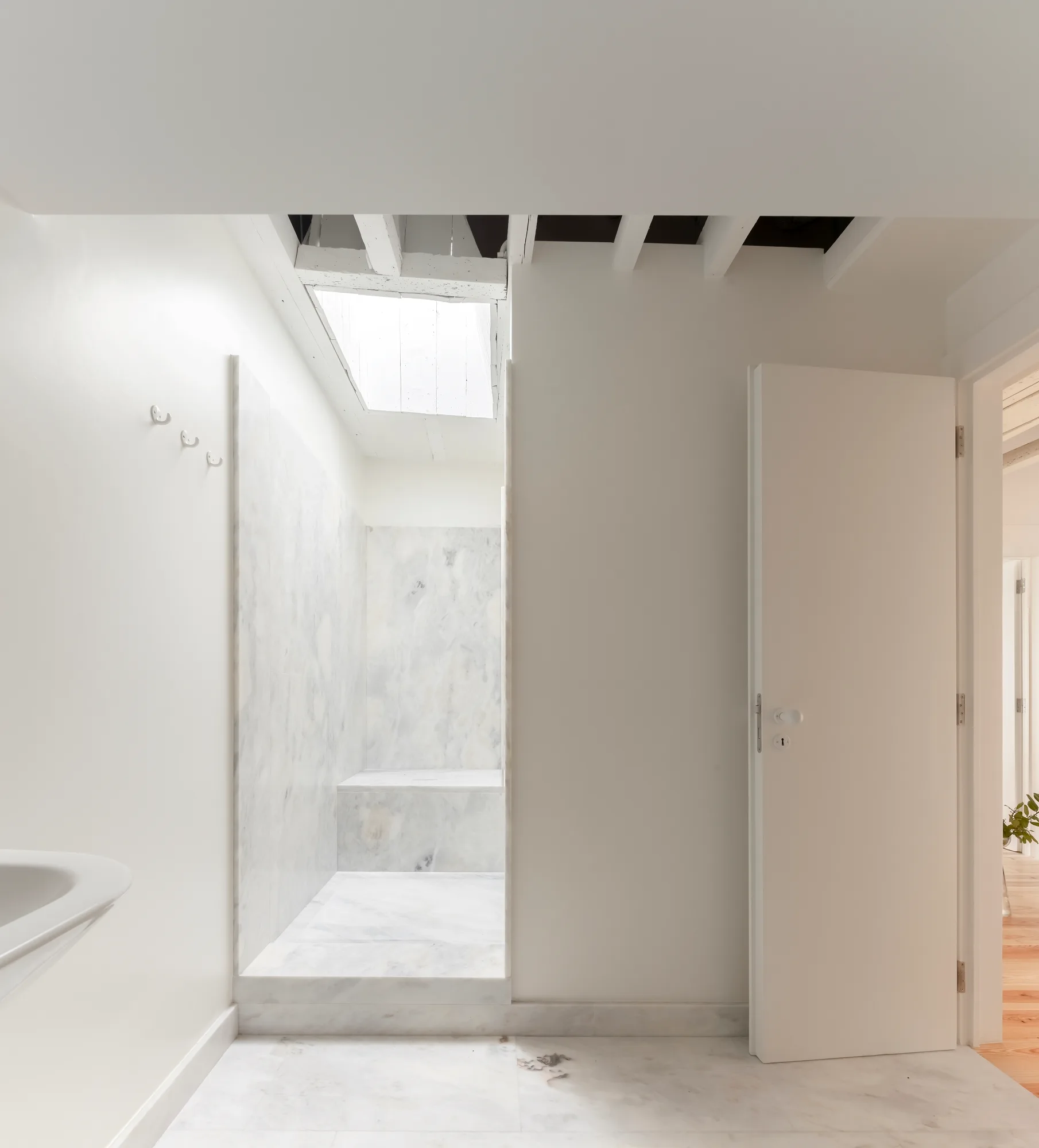 A minimalist white bathroom features a marble-tiled shower stall positioned beneath an open ceiling skylight next to an open white door.
