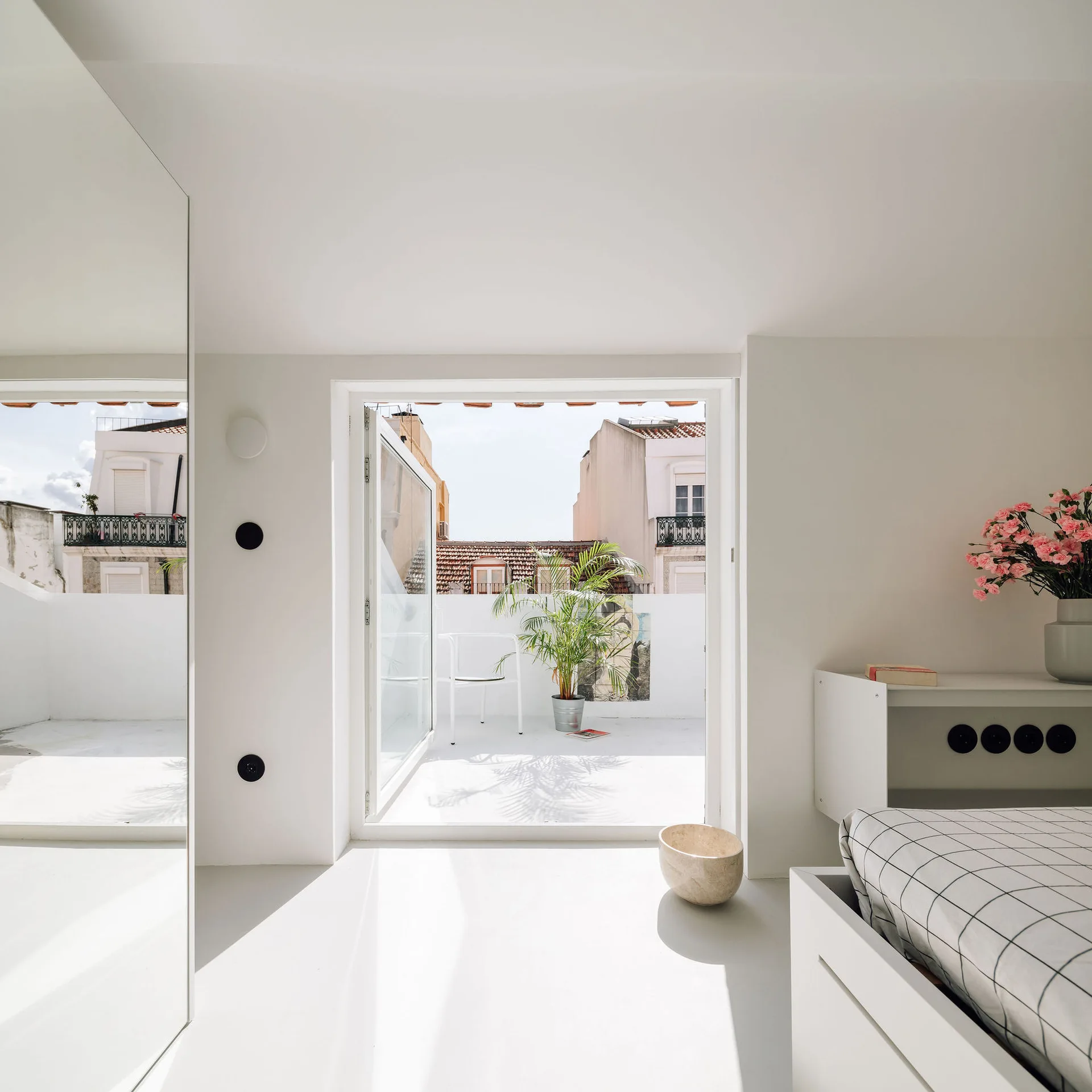 A minimalist white bedroom features a grid-patterned bed, a large floor mirror, and an open door leading to a sunny balcony with a potted plant.