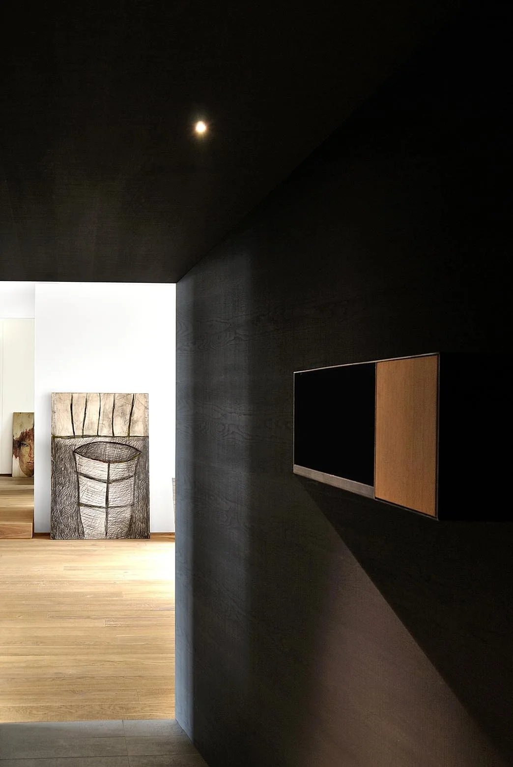A dark wood paneled hallway with a floating cabinet looks into a bright room with light wood floors and a large sketch.