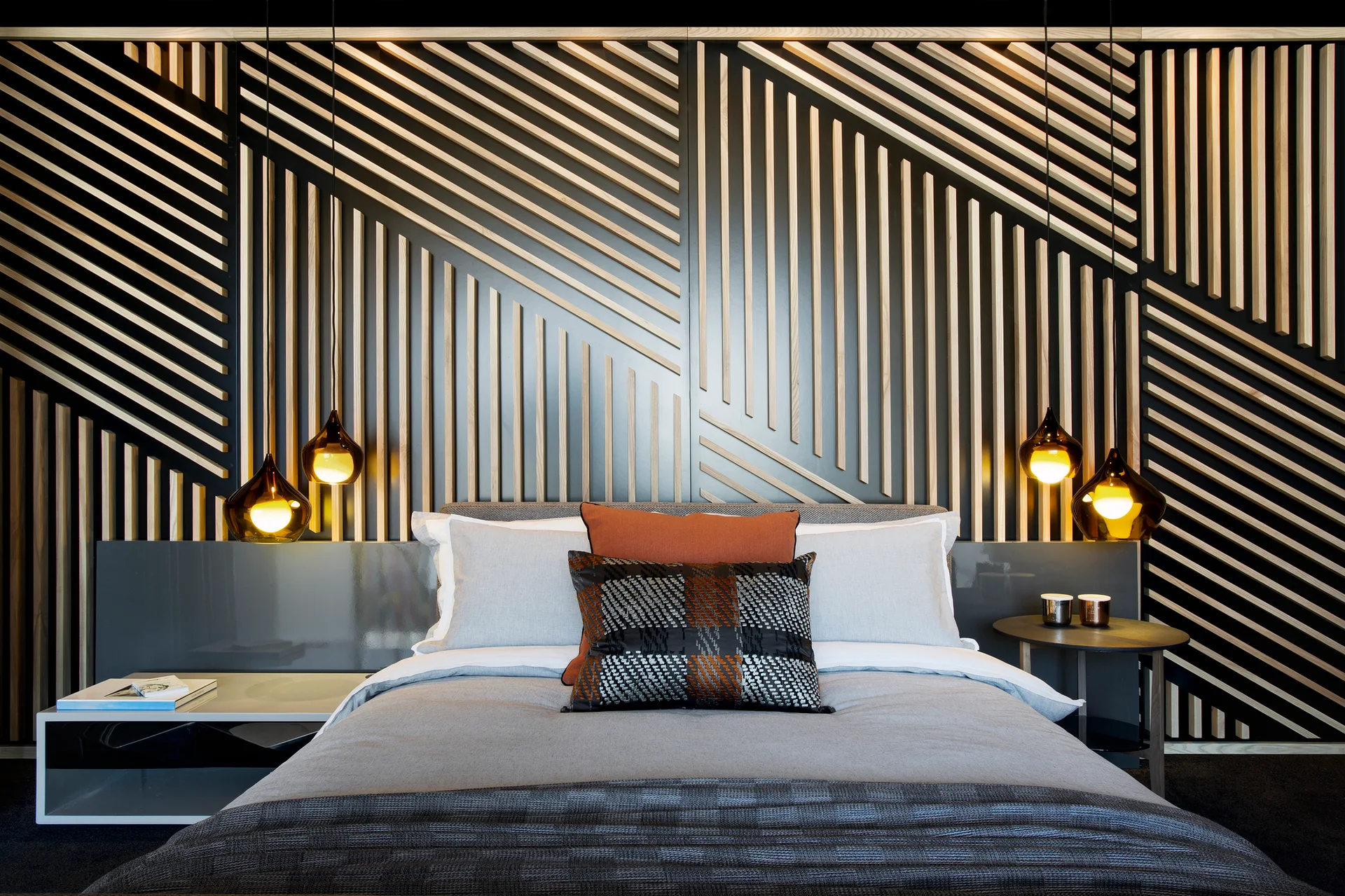 A modern bed with grey and rust pillows is set against a geometric black and wood slat accent wall with amber glass pendant lights.
