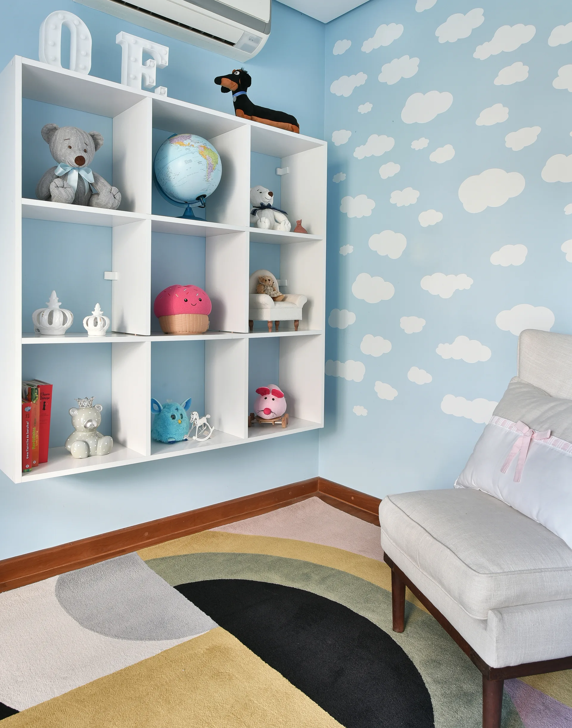 A children's room features a white grid wall shelf filled with toys next to light blue cloud wallpaper and a colorful geometric rug.