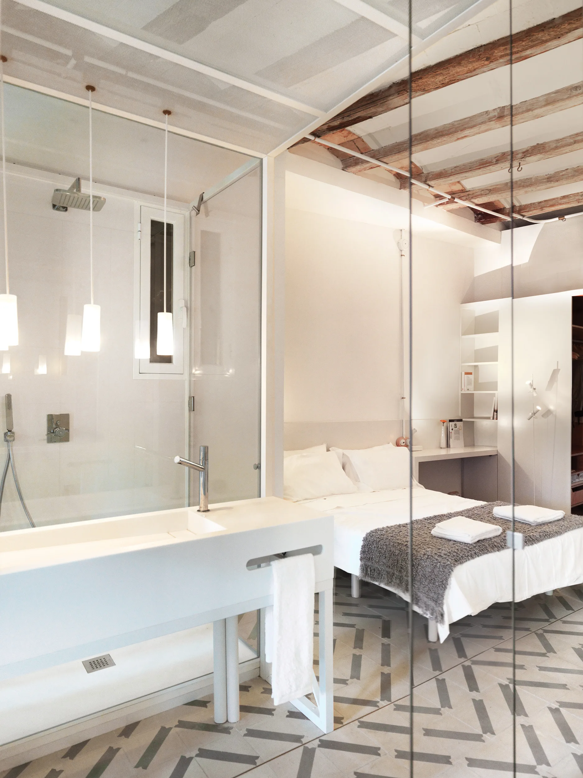 An open-plan bedroom and bathroom suite features geometric floor tiles, a glass partition, a minimalist white sink, and exposed wooden ceiling beams.