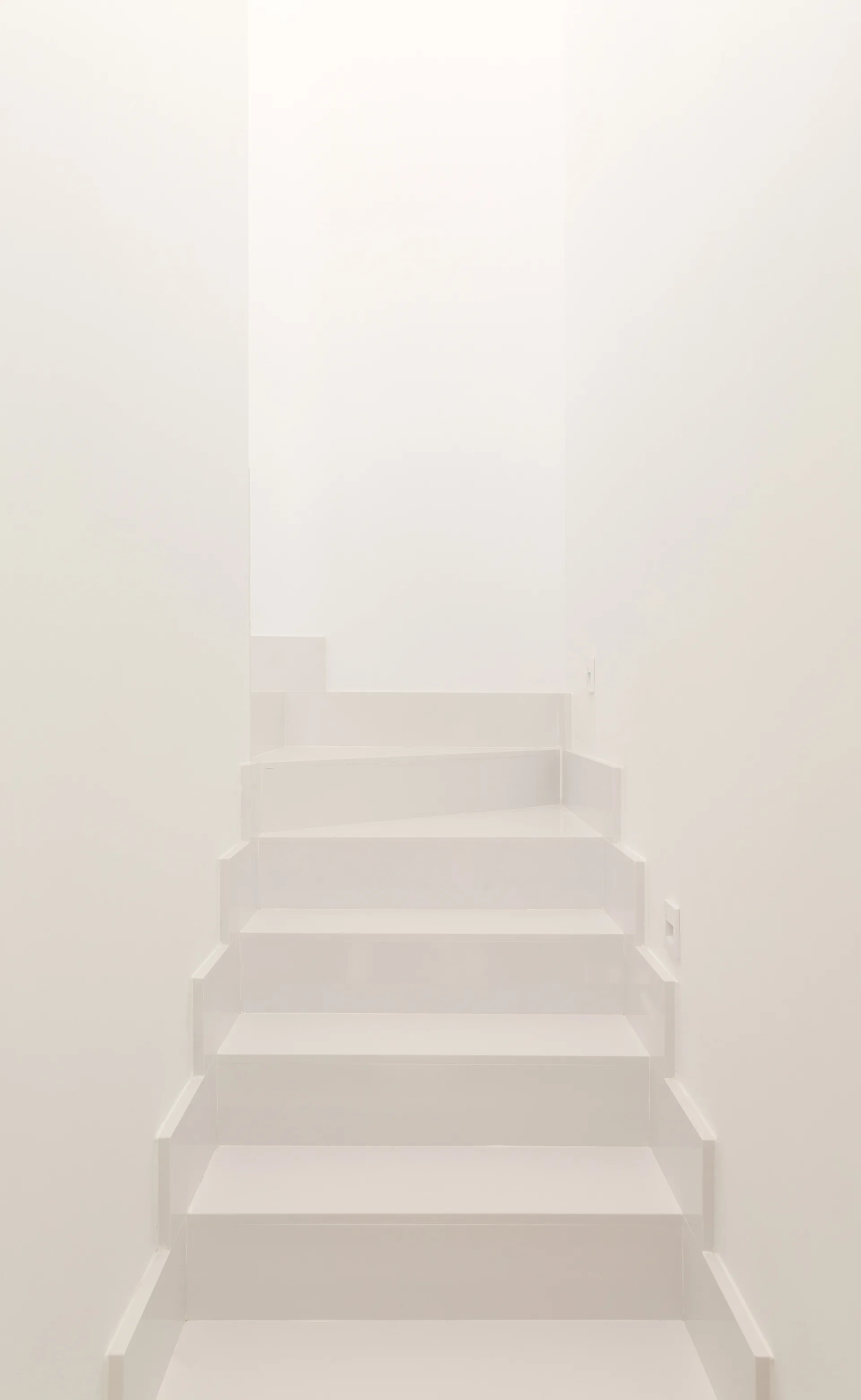 A minimalist white staircase ascends between smooth white walls in a monochromatic interior stairwell.