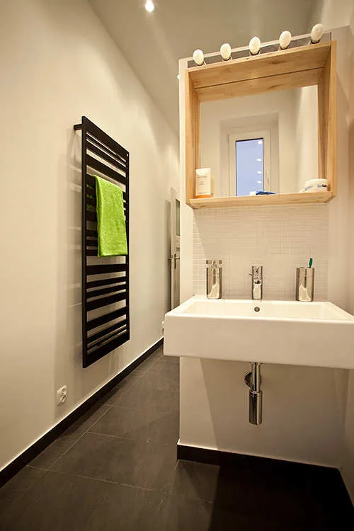 A narrow modern bathroom with a white wall-mounted sink, a wood-framed mirror with light bulbs, and a black wall towel rack.