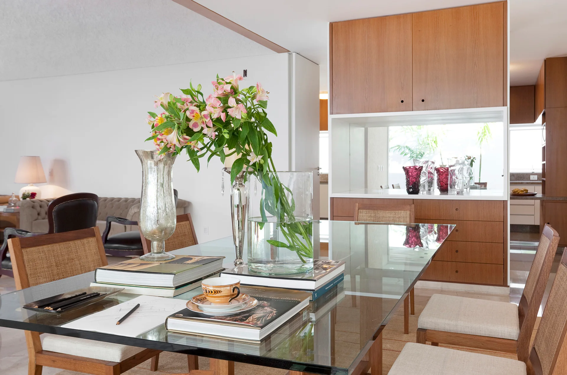 A glass-top dining table surrounded by cane-back chairs is styled with stacked books and flowers in front of a warm wood cabinet.