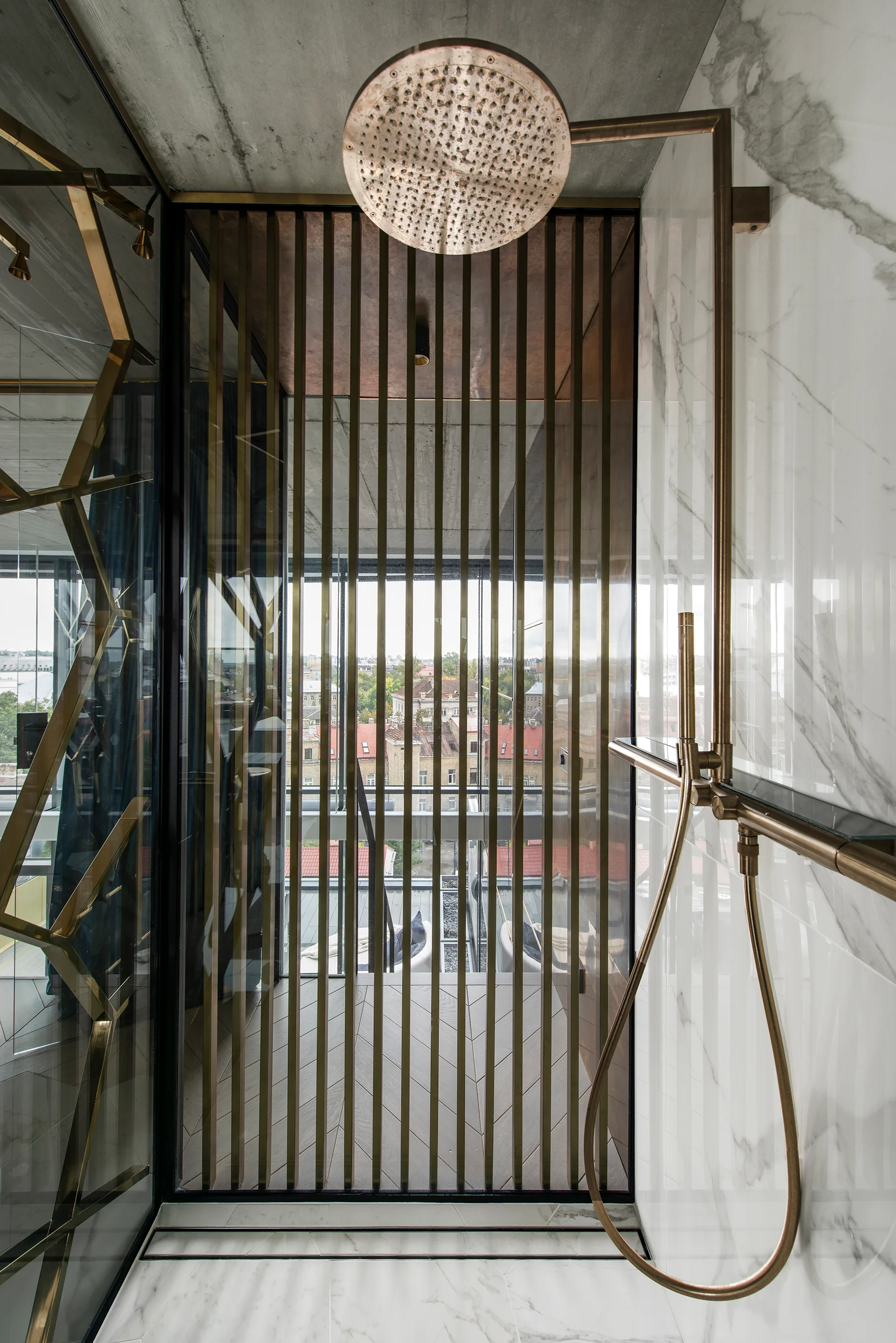 A modern shower features white veined marble tile, a bronze rainfall shower head, a raw concrete ceiling, and a vertical bronze metal slatted partition.