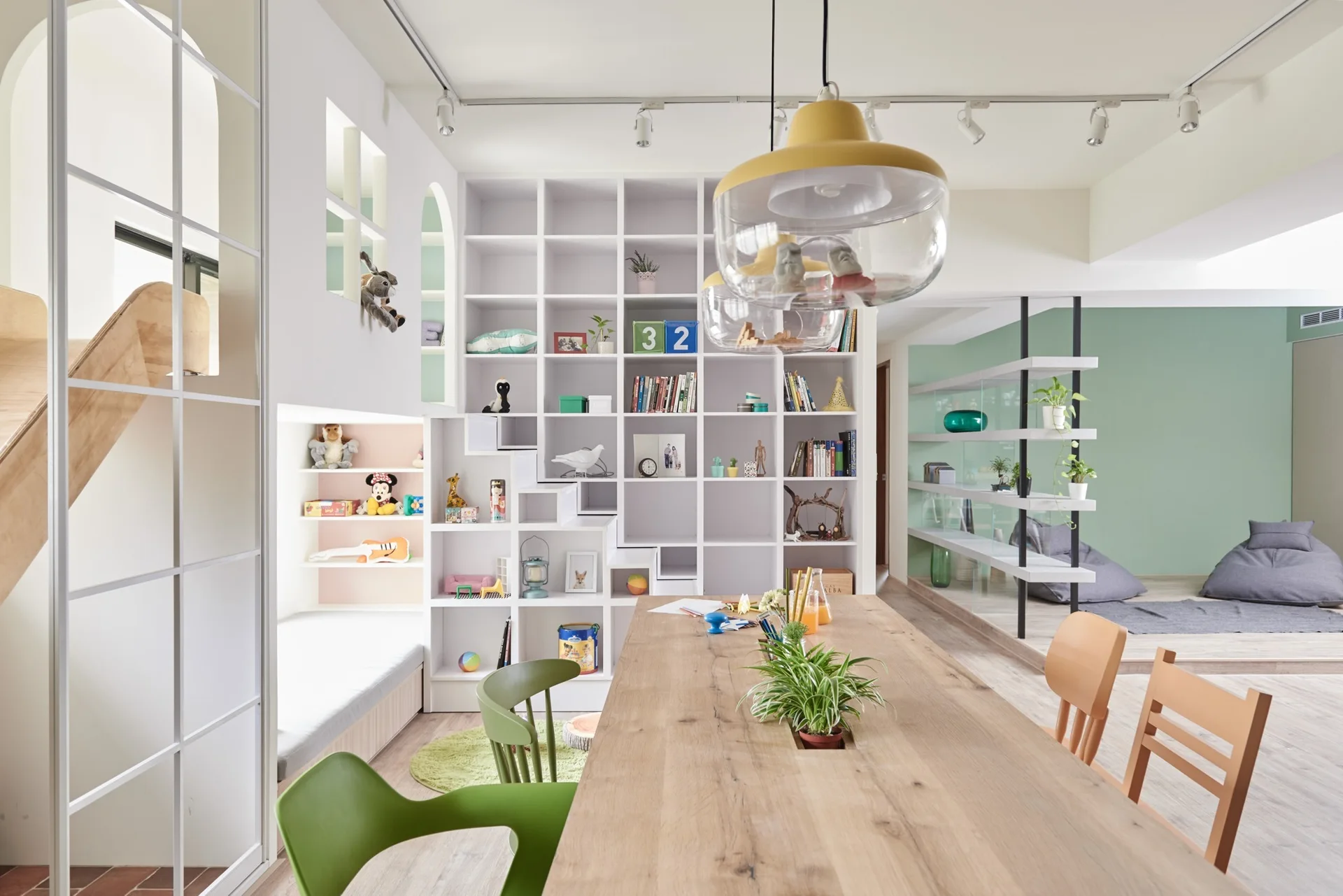 A bright playroom features a long wooden table with green chairs, yellow pendant lights, and a large white gridded shelving unit with a built-in nook.