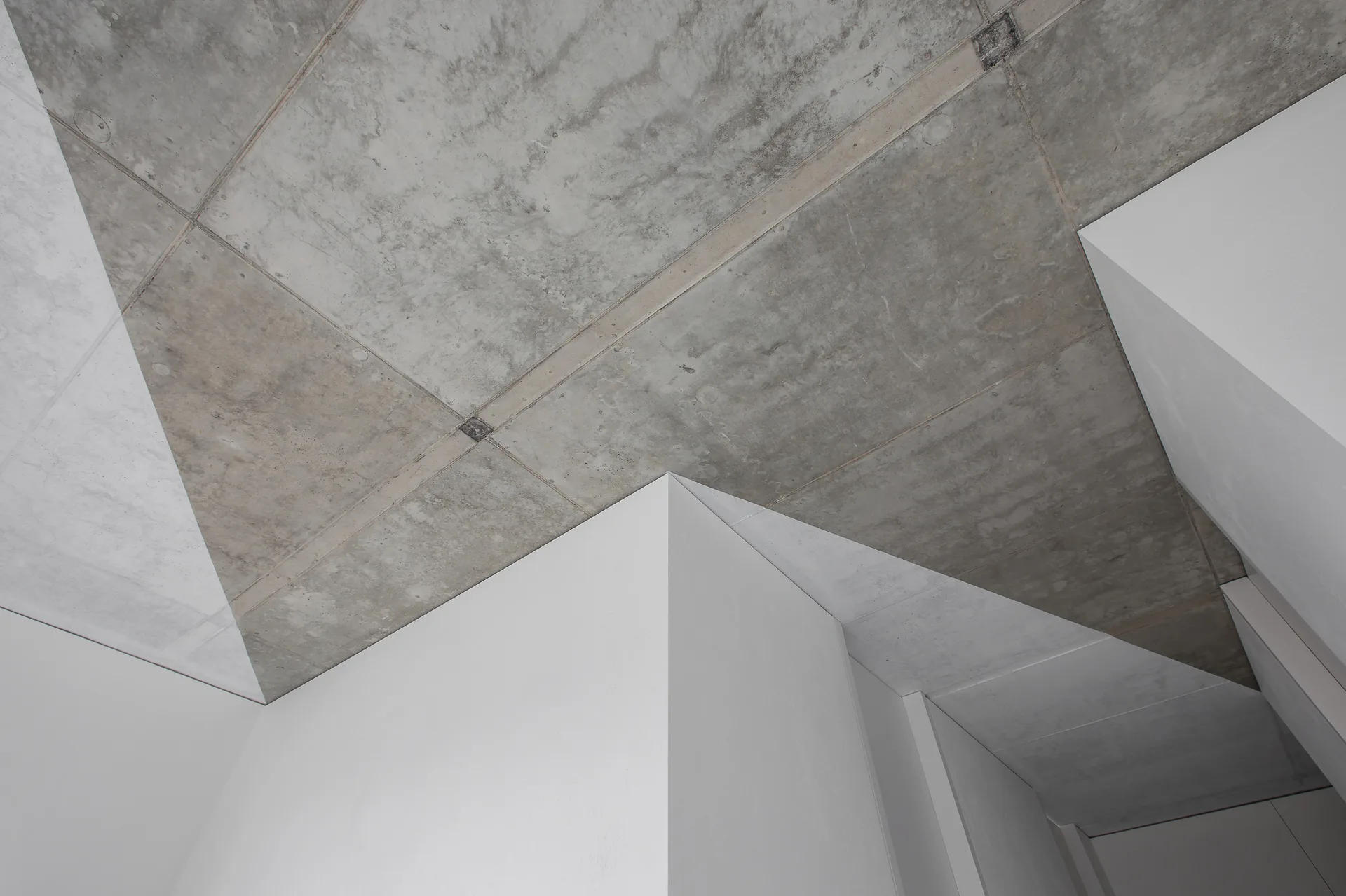 A close-up of a raw grey concrete ceiling above sharp geometric white drywall structures in a minimalist interior.