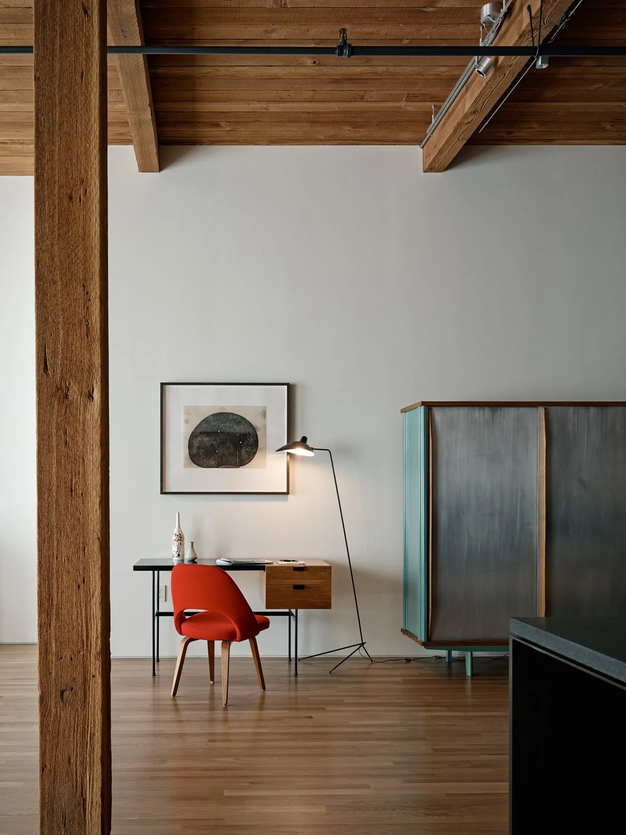 A minimalist home office workspace features a light wood desk, a red chair, a slim floor lamp, and exposed timber ceiling beams.