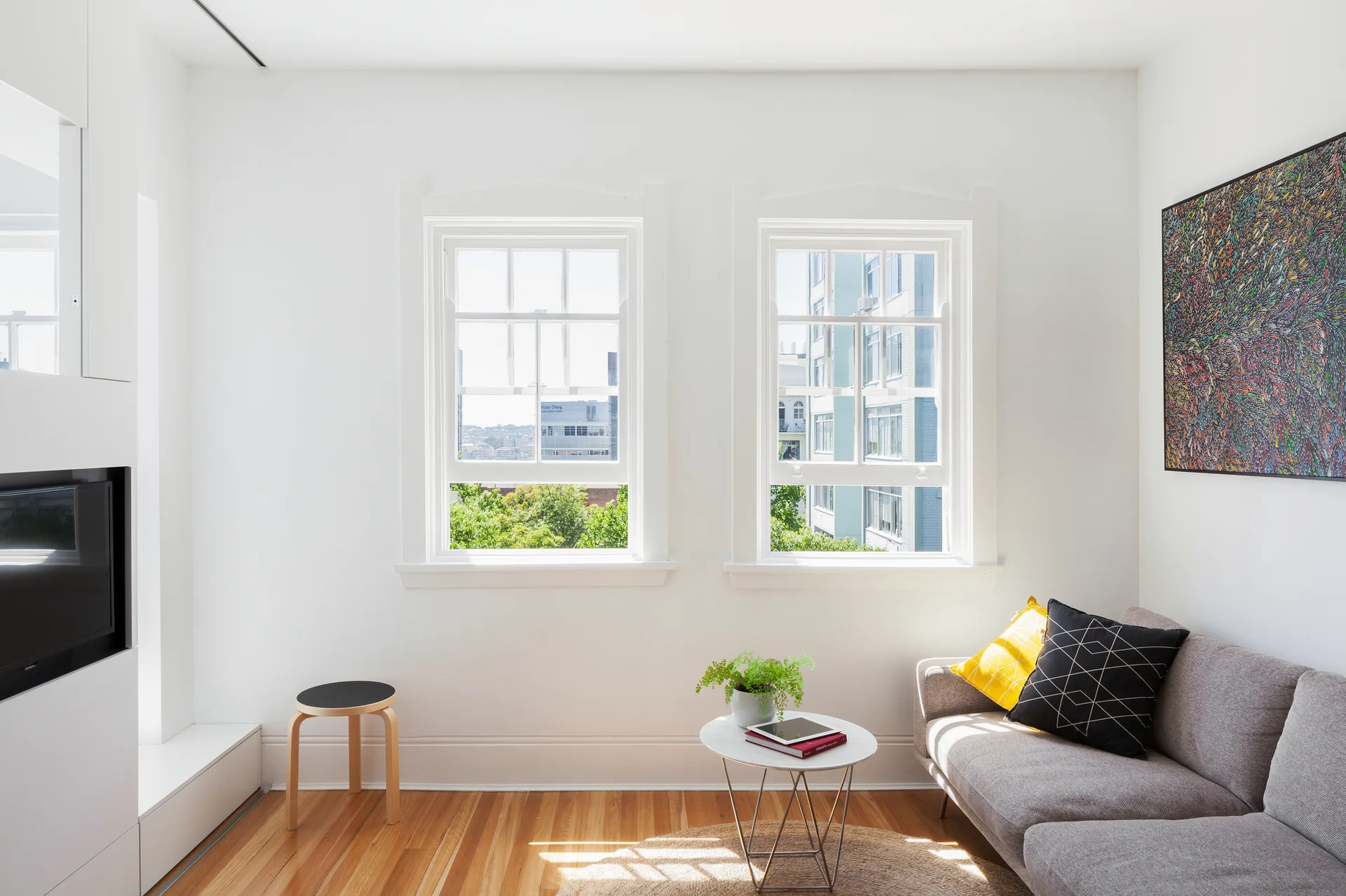 A bright white living room featuring a gray fabric sofa with yellow and black pillows, two sash windows, and warm wood floors.