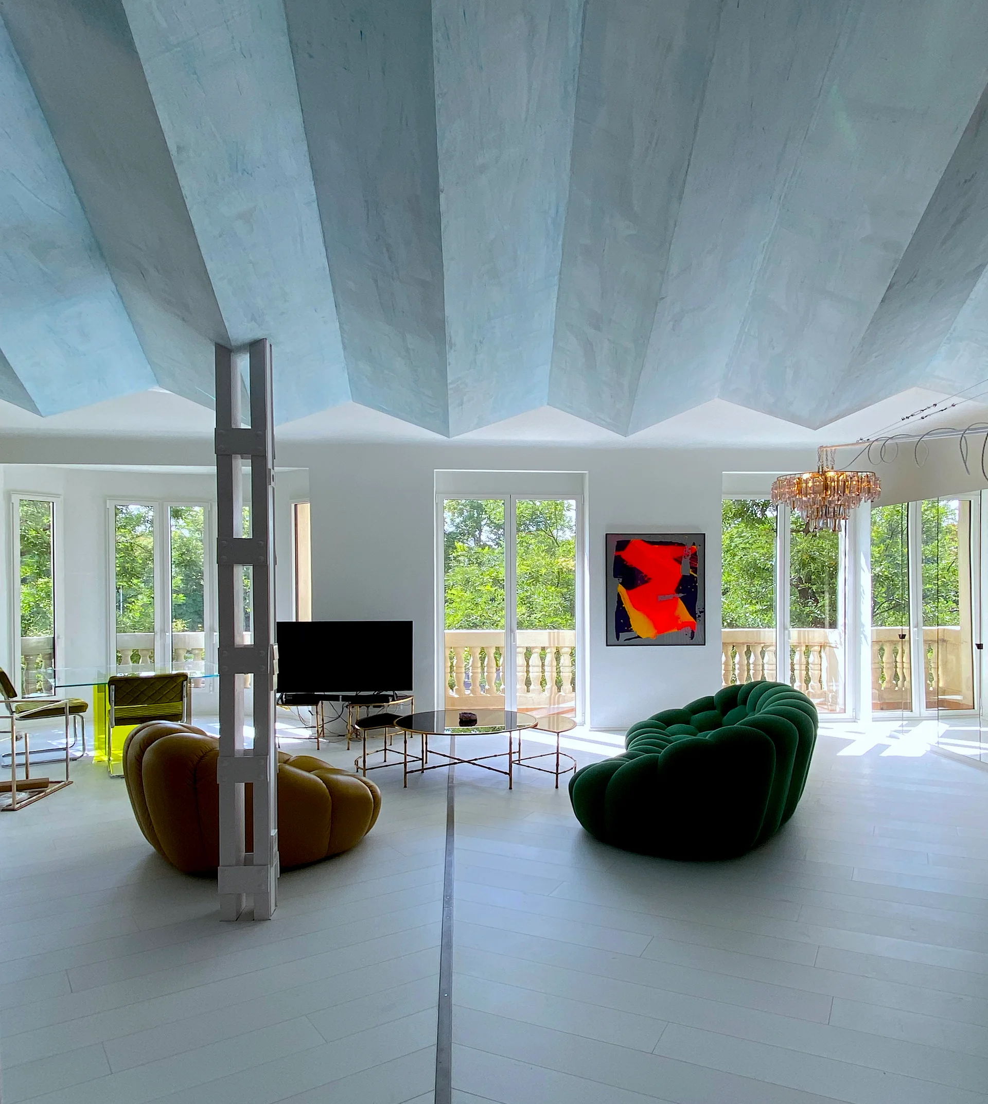 A bright modern living room featuring a light blue pleated ceiling, curved green and brown sofas, and tall windows.