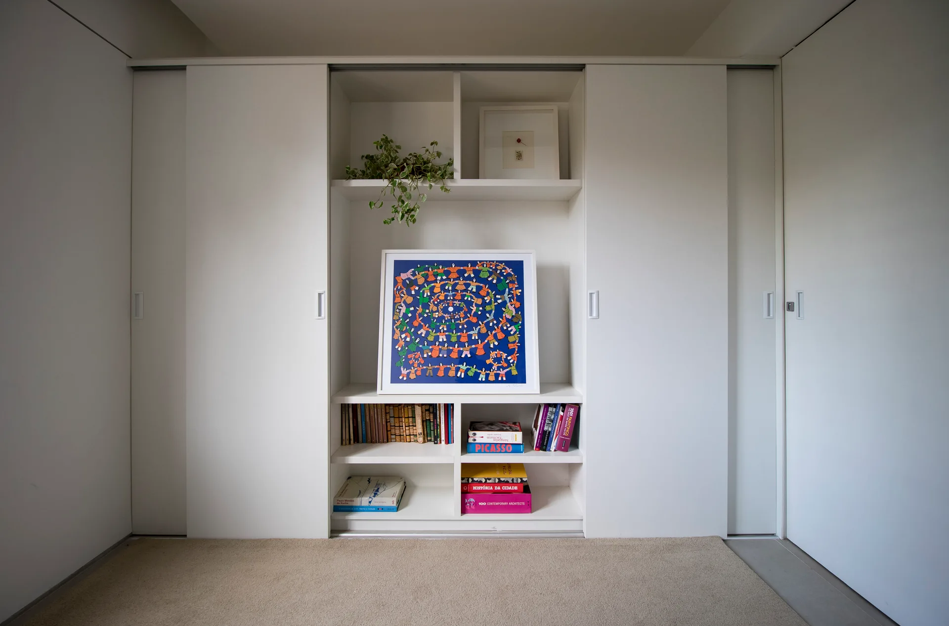White built-in closet with sliding doors featuring central open shelving that displays books, a trailing plant, and a framed colorful art print.