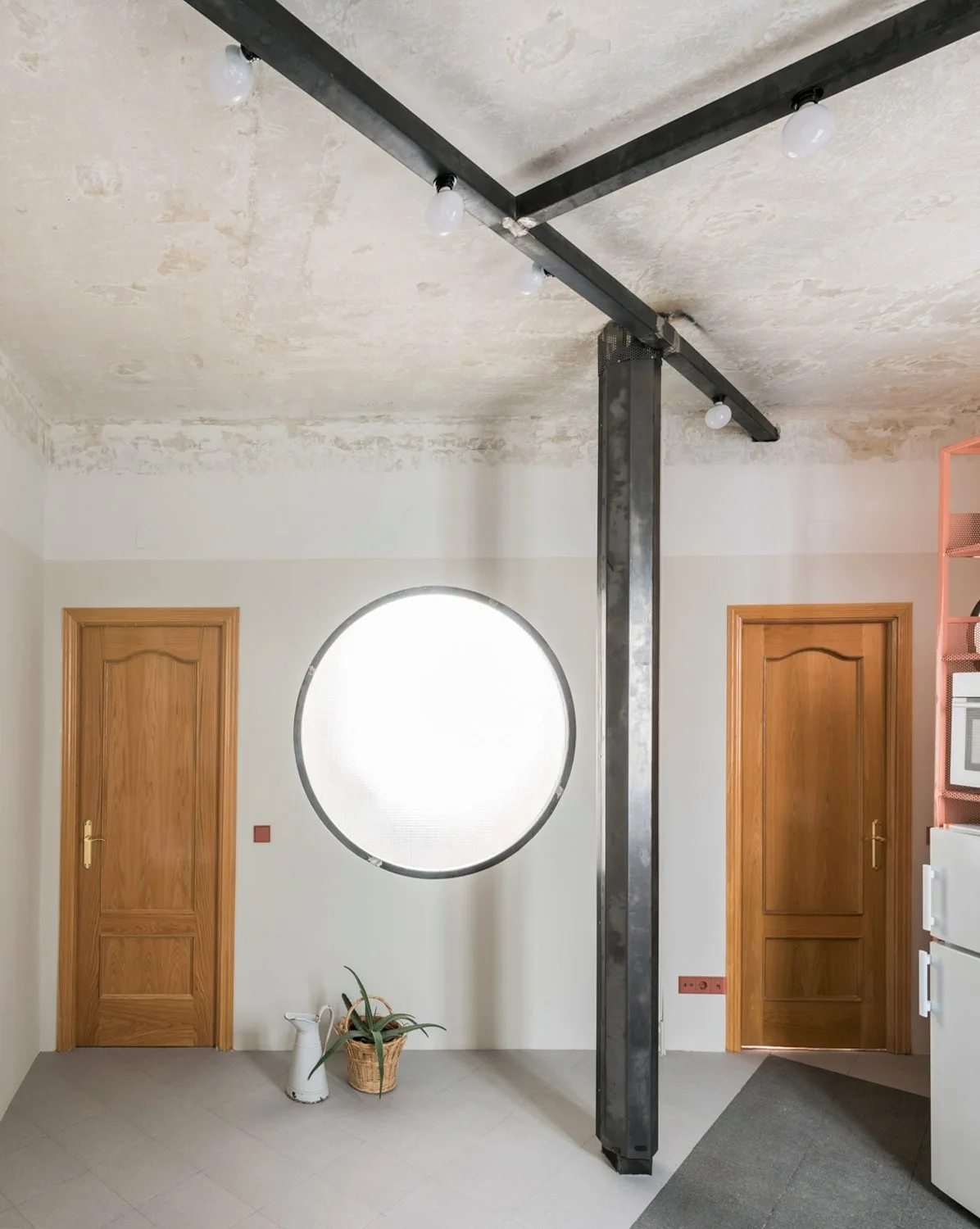 A modern hallway features two wooden doors flanking a circular frosted window under an exposed steel beam and concrete ceiling.