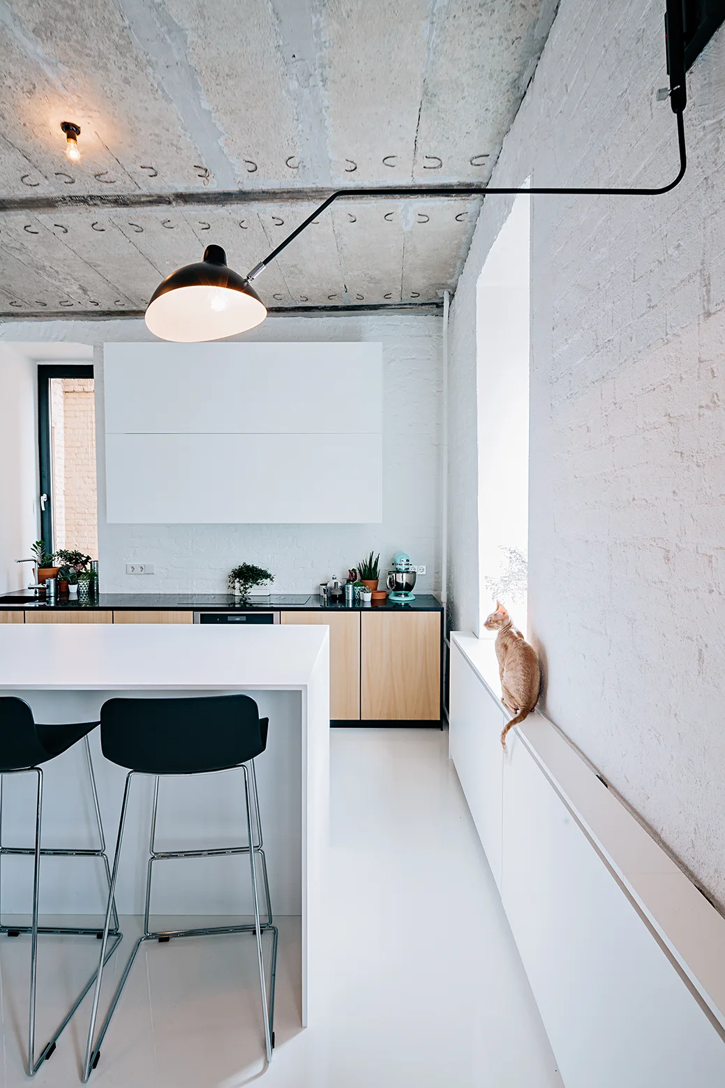 A minimalist kitchen with a white island, black bar stools, light wood cabinets, white brick walls, and an exposed raw concrete ceiling.