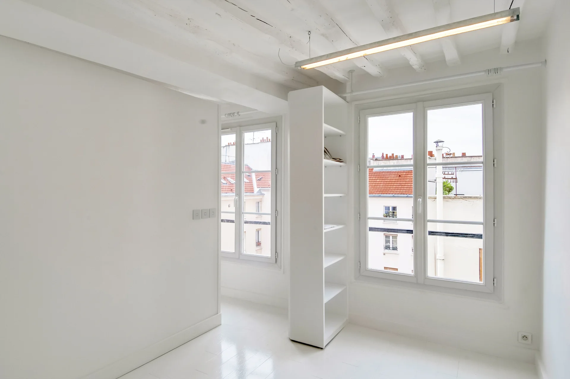 An empty white room features painted exposed ceiling beams, a tall white shelving unit divider, and two multi-pane windows showing rooftops.