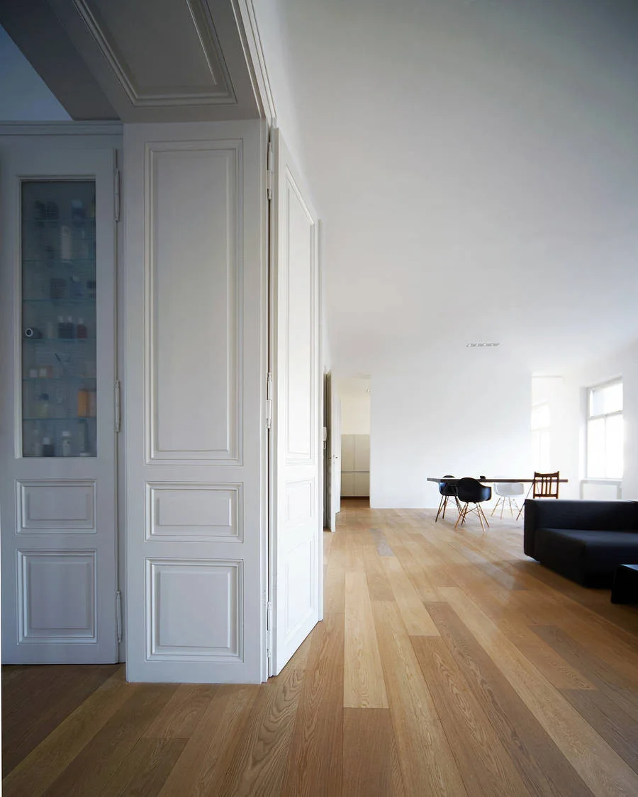 A bright open-concept living space features light oak plank flooring, a white paneled doorway with built-in glass storage, and a minimalist dining area.