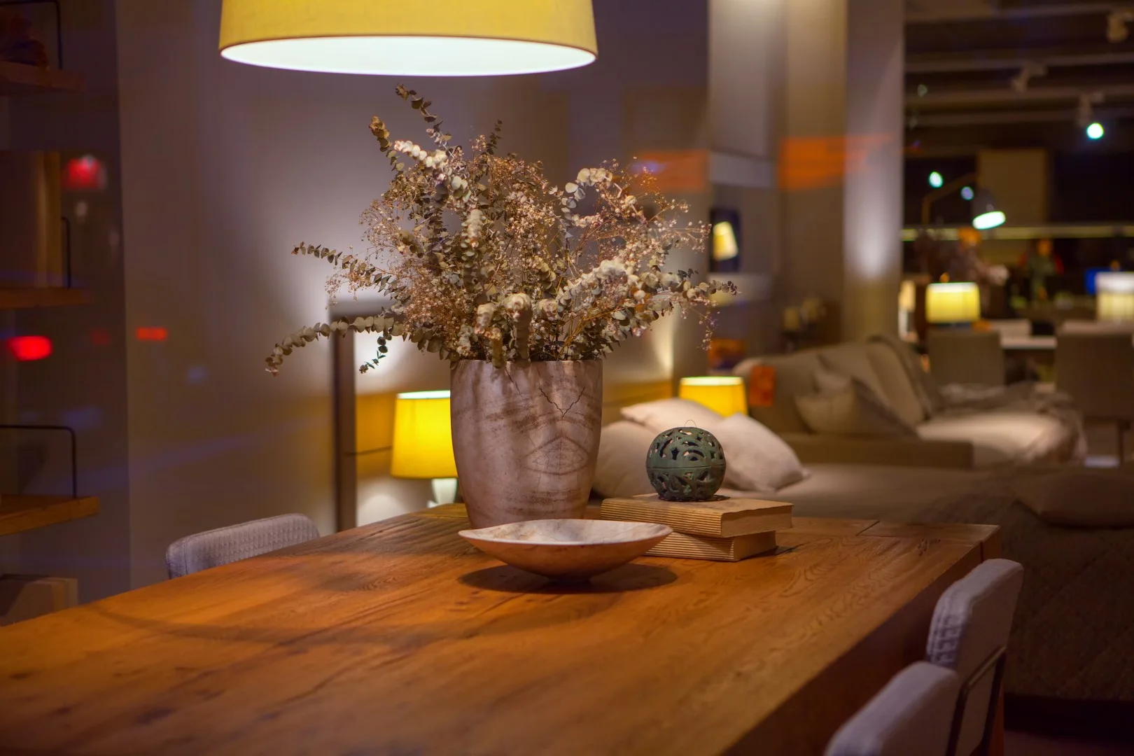 A rustic wood dining table features a centerpiece of dried eucalyptus branches in a textured vase under a warm pendant light.