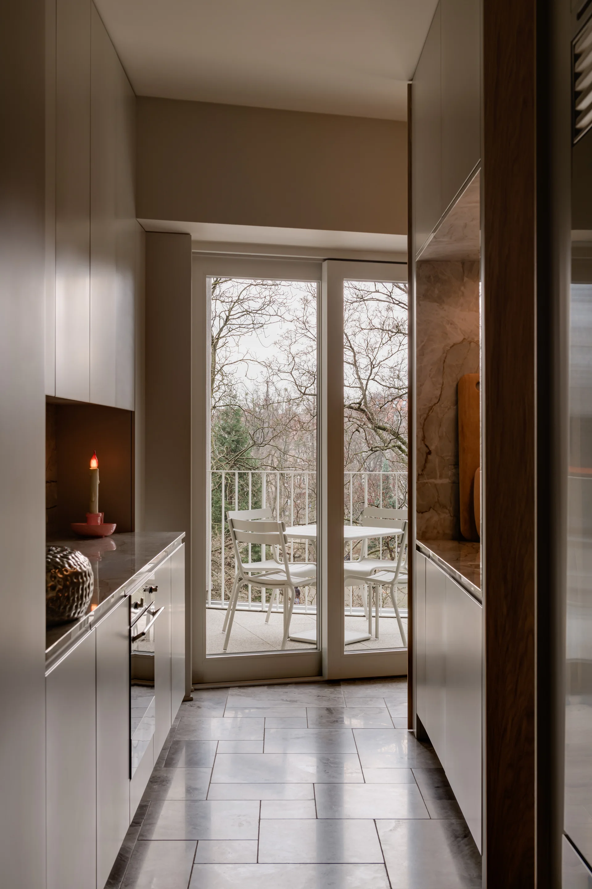 A narrow galley kitchen with taupe cabinets and stone countertops leads to a glass door looking out to a balcony with white patio chairs.