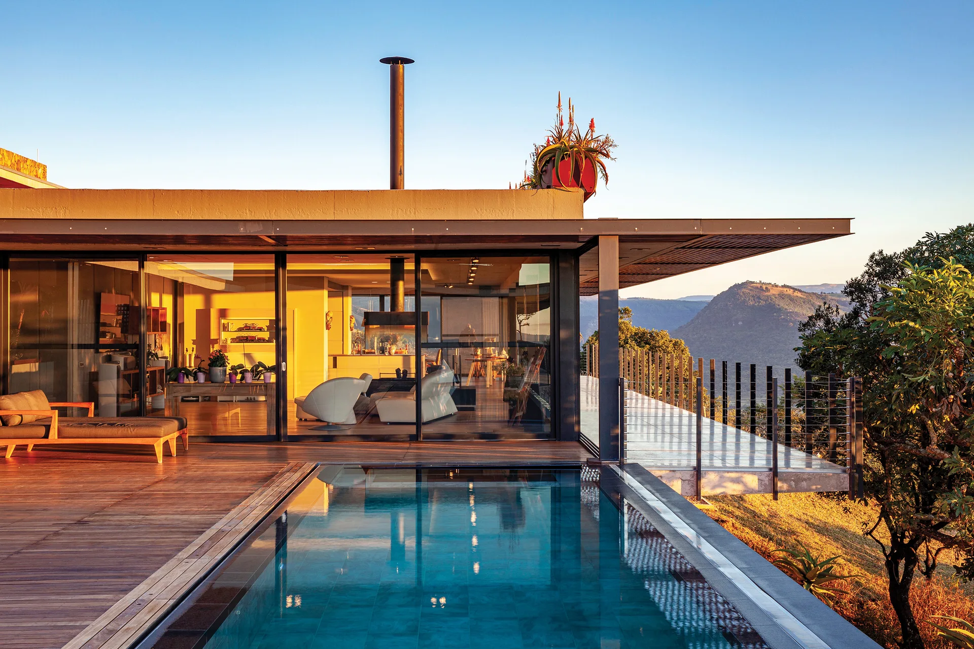 A modern hillside home features a rectangular swimming pool surrounded by a wooden deck, with floor-to-ceiling glass walls showing the interior at sunset.