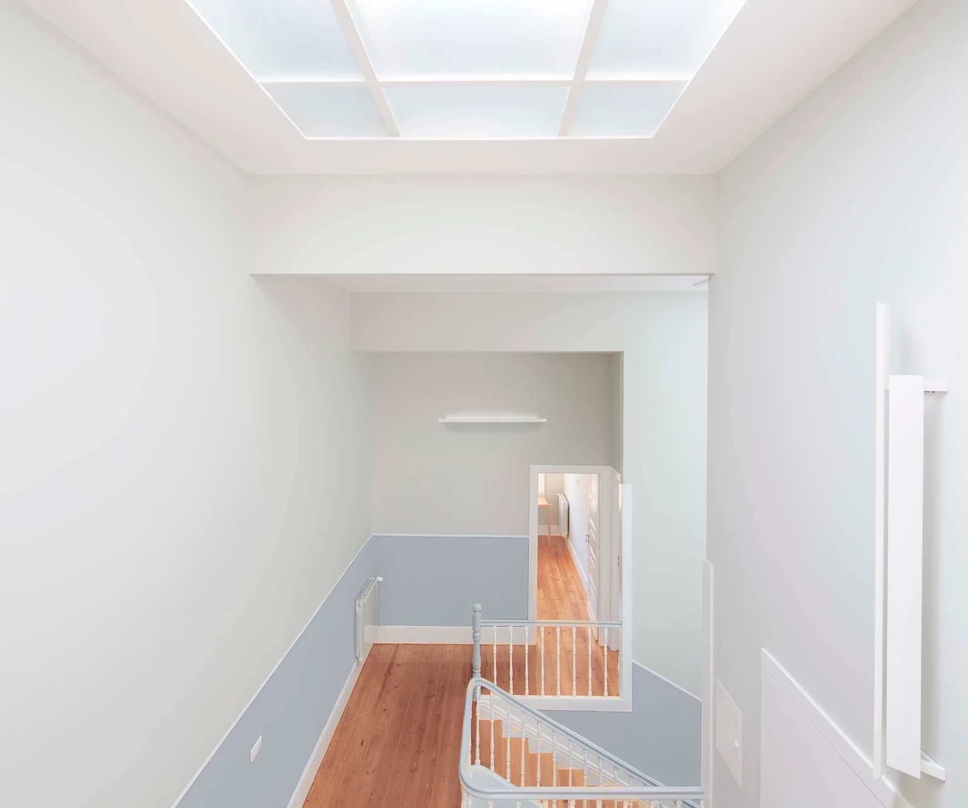 A high-angle view looking down a wooden staircase with a grey railing, two-tone blue and white walls, and a large grid-paned skylight overhead.