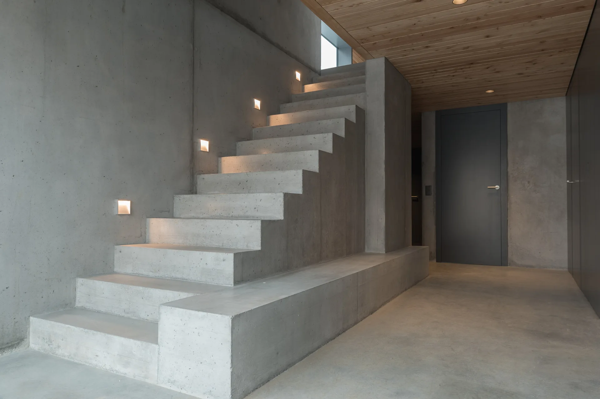 A minimalist concrete staircase rises alongside a concrete wall with square recessed lights under a warm wood plank ceiling.