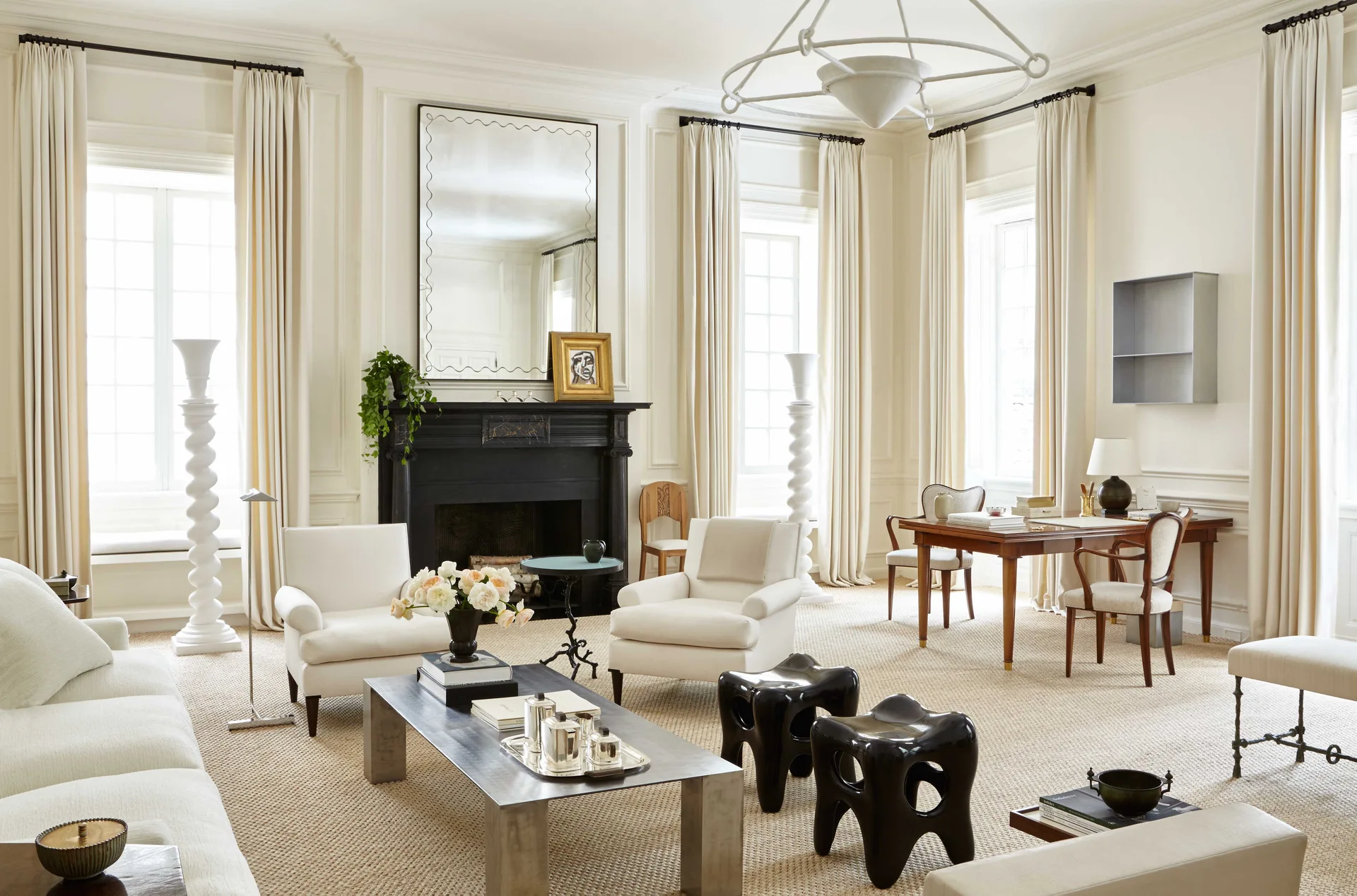A bright cream-colored living room featuring a black fireplace mantel, white upholstered chairs, tall sculptural floor lamps, and a wooden writing desk.
