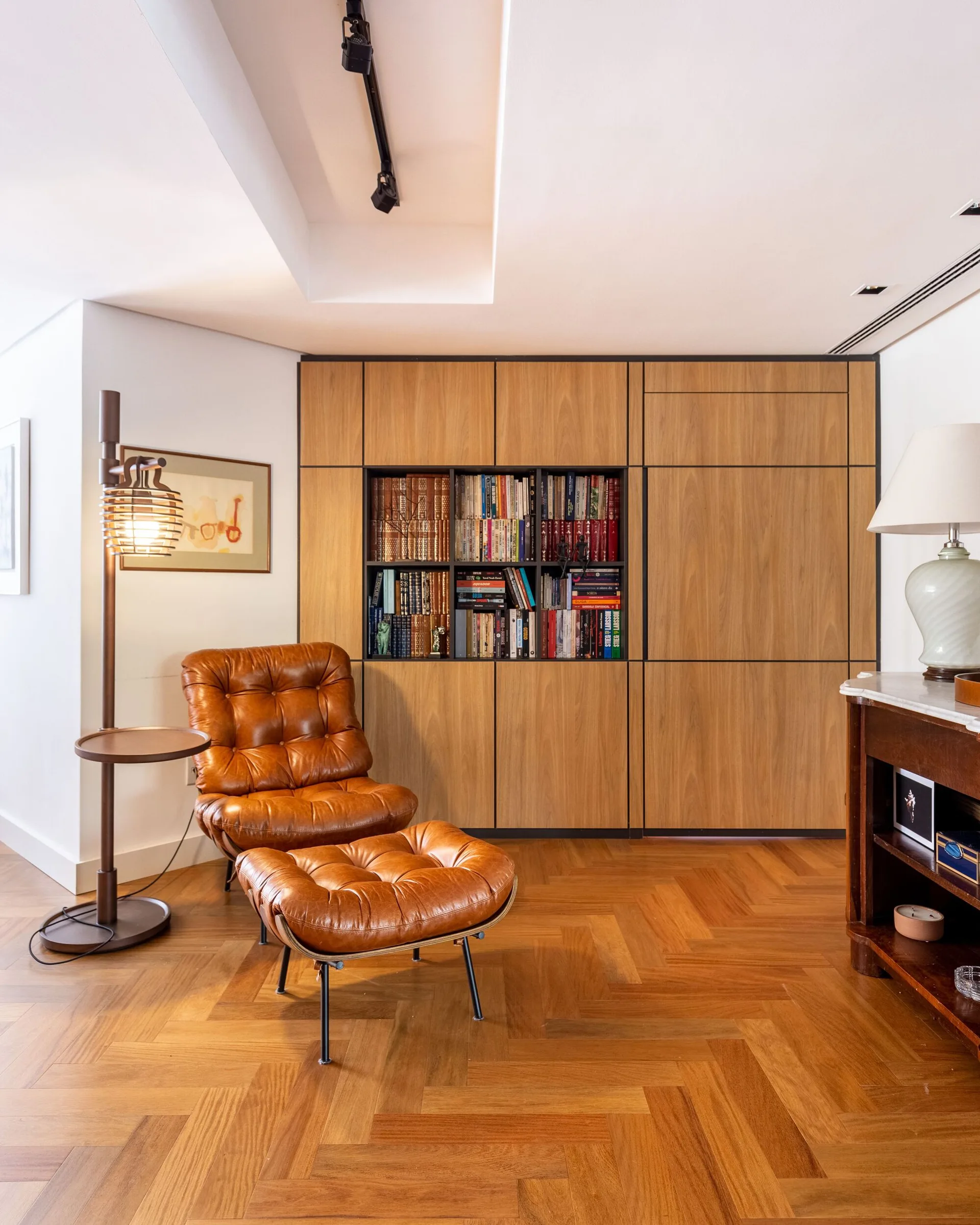 A brown tufted leather lounge chair and ottoman sit on a herringbone wood floor in front of a flat-panel wood storage wall with open shelving.