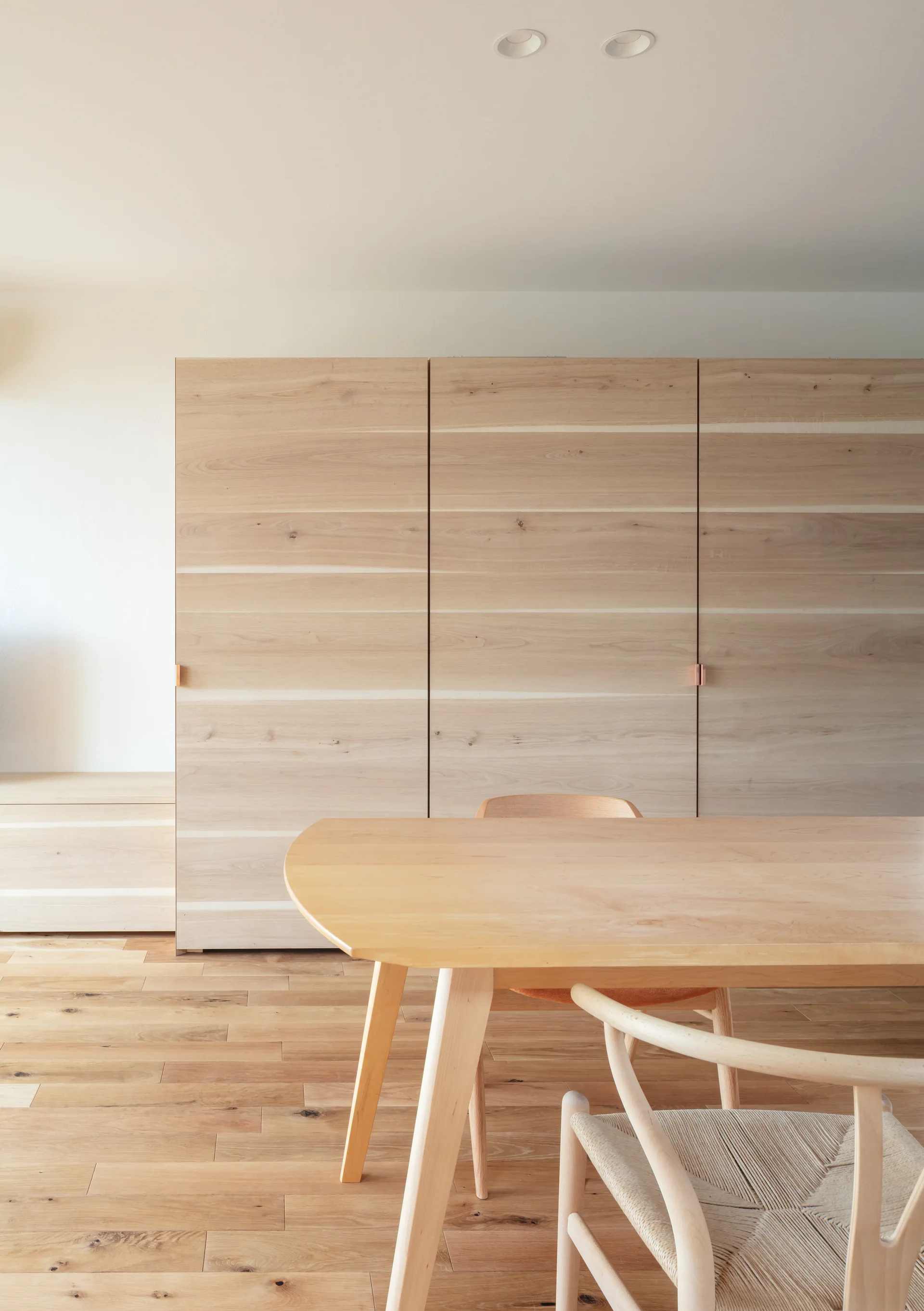 A minimalist dining room features a light wood table, a woven-seat chair, and large, flat-front wooden storage cabinets on wood floors.