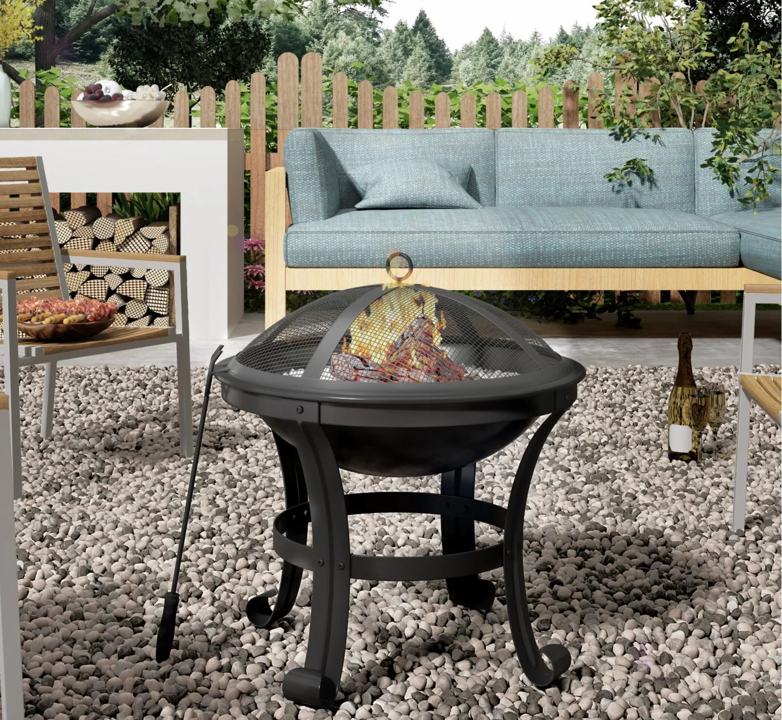 A black metal fire pit burns on a gravel patio next to a blue patterned outdoor sectional sofa and a wooden picket fence.