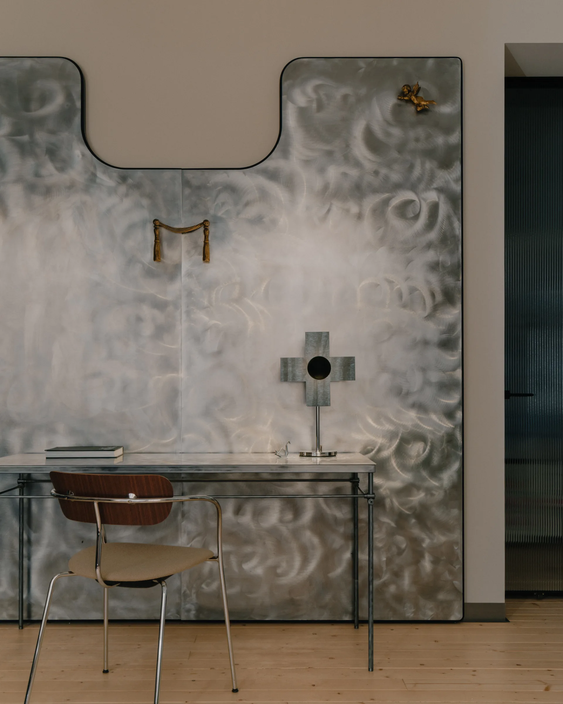 A modern writing desk and wood-backed chair are positioned against a large brushed-metal wall panel with a swirling texture.