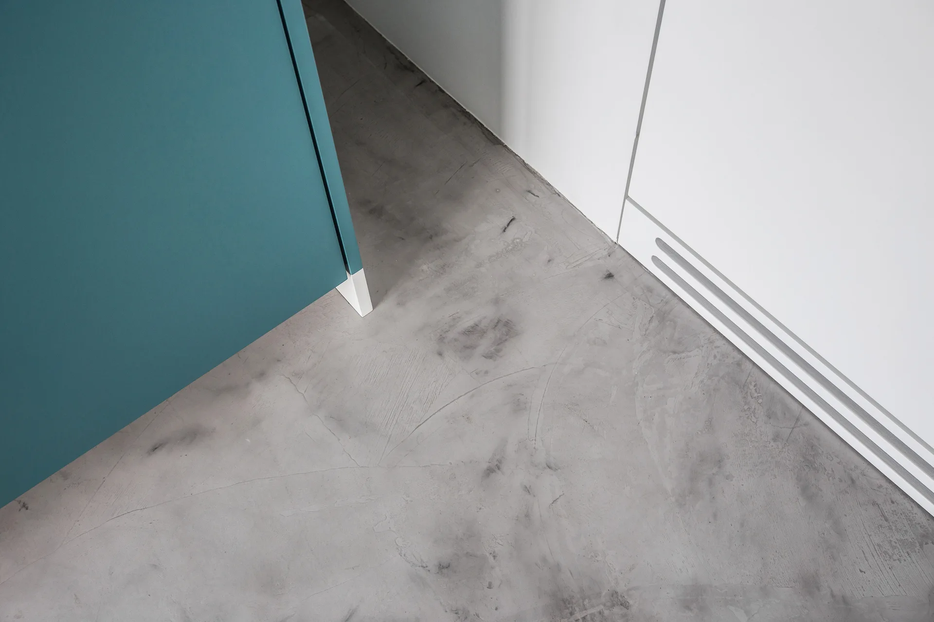 A high-angle view of a teal cabinet corner and a white vent paneled base meeting textured gray concrete floors.