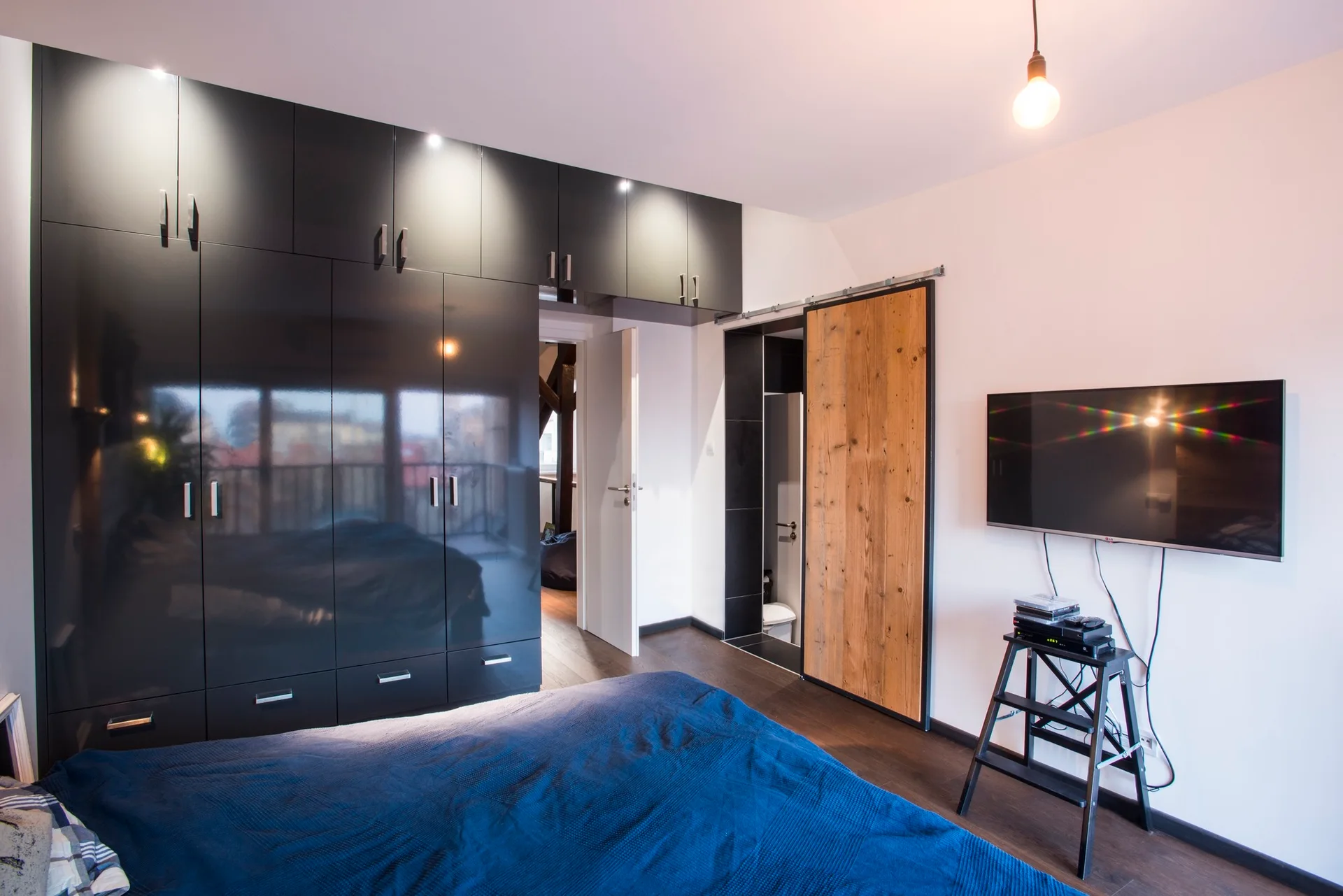 A modern bedroom featuring high-gloss black built-in wardrobes, a sliding wooden barn door, and a bed with a blue cover.