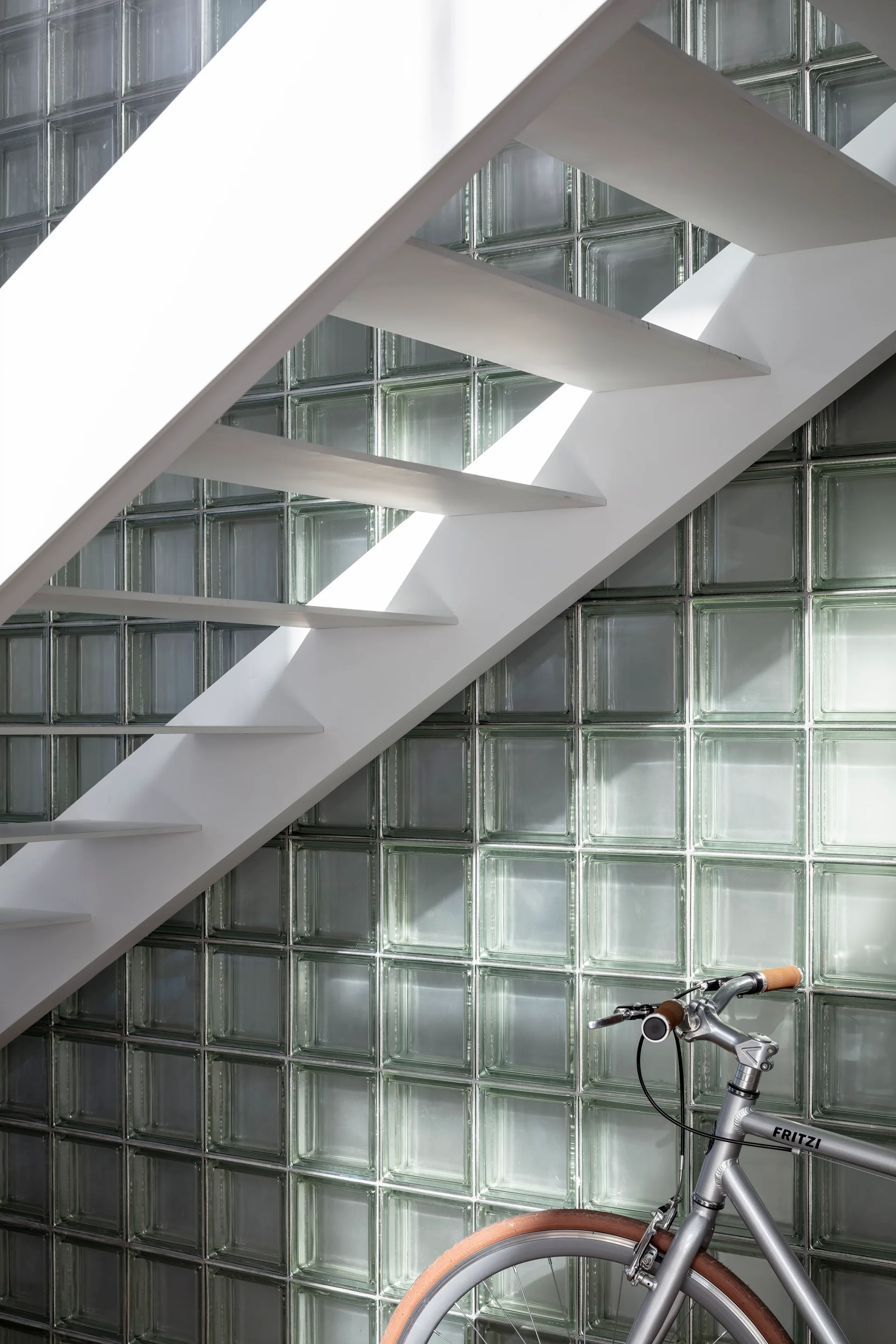 A white open-tread staircase climbs diagonally in front of a translucent glass block wall, with a silver bicycle partially visible below.