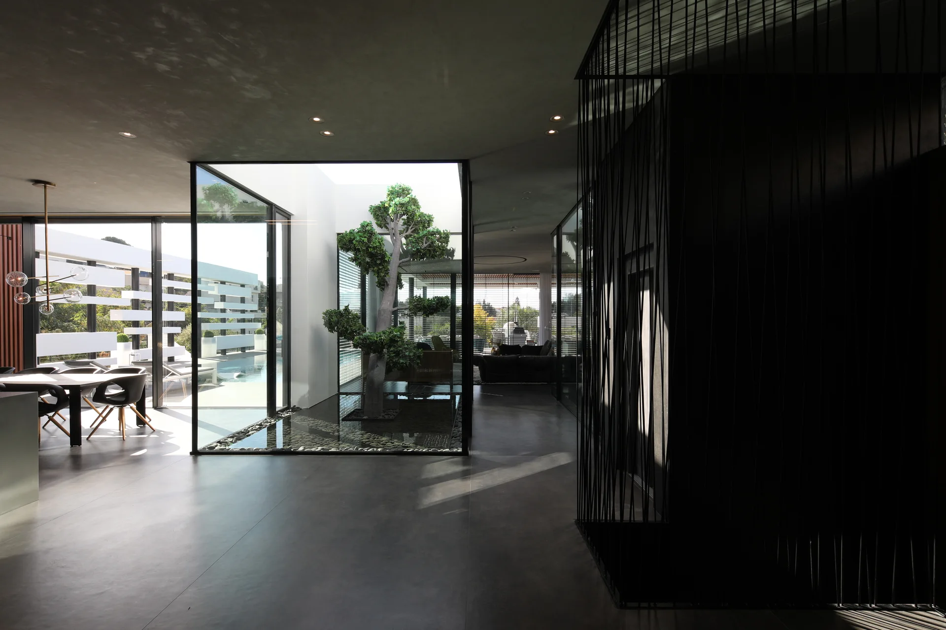 A dark minimalist interior features a glass-enclosed central courtyard with a green tree on a pebble bed next to a dining area.