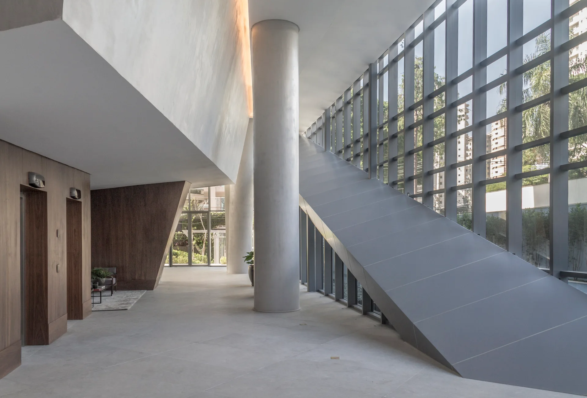 A spacious modernist lobby featuring an angular grey staircase, a large cylindrical concrete column, wood-paneled walls, and a glass grid curtain wall.